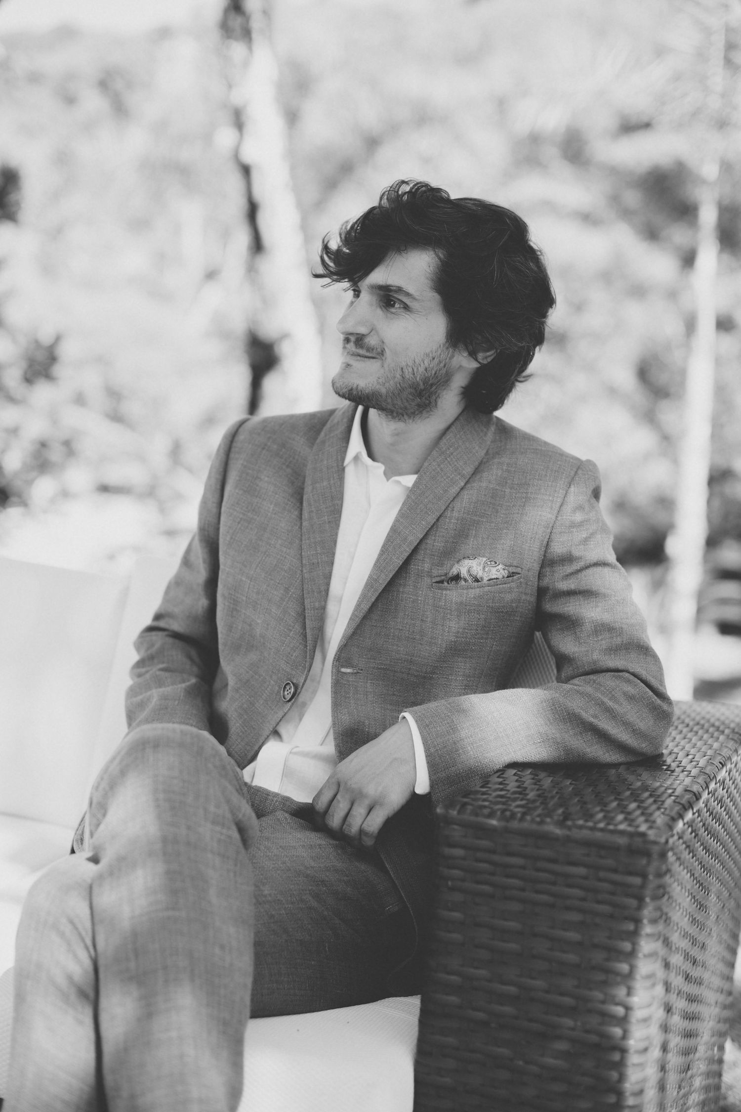 A black and white portrait of someone in a suit sitting on wicker furniture.