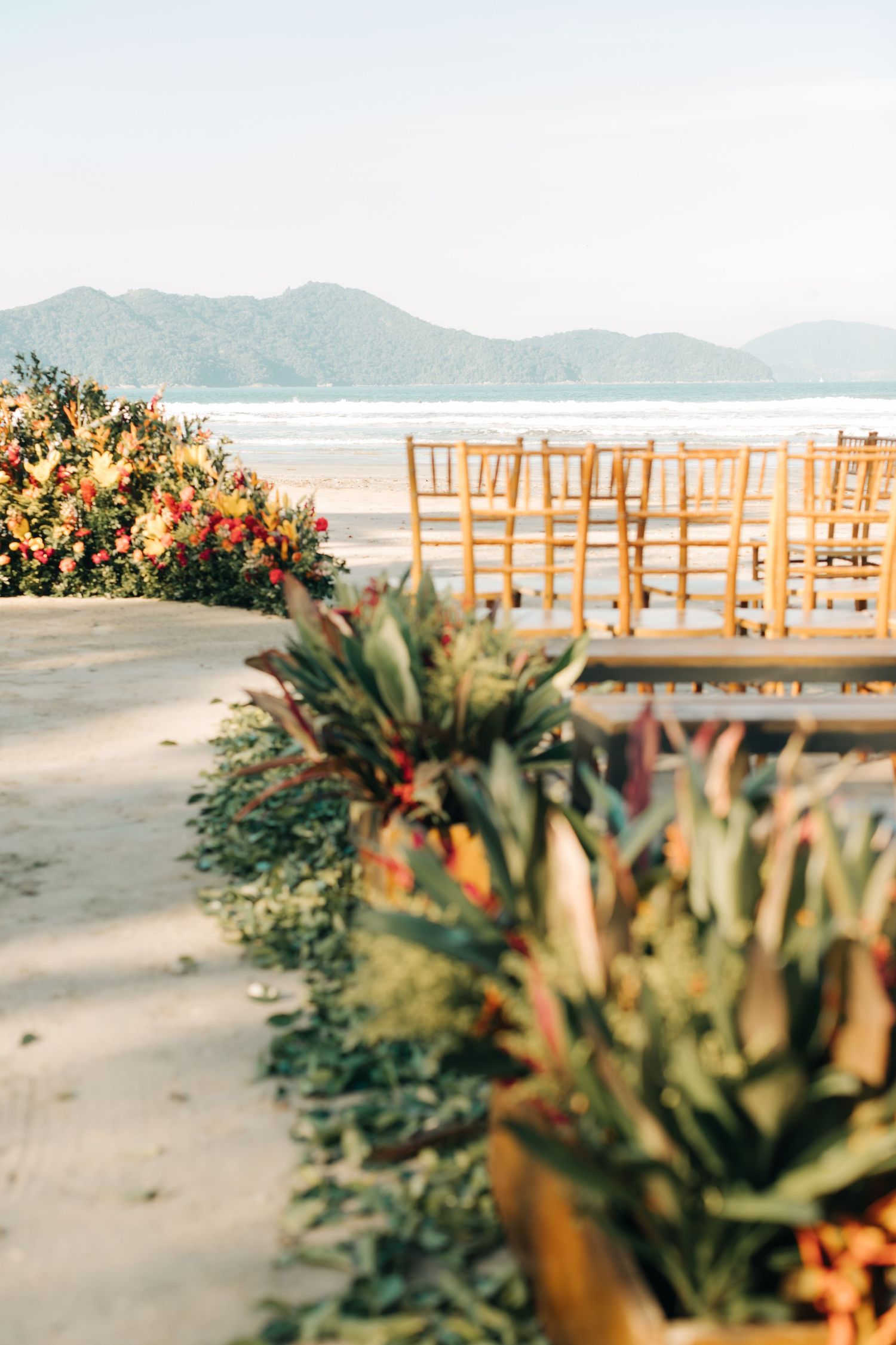 Wooden chairs and tropical flowers line a beachside wedding ceremony aisle.