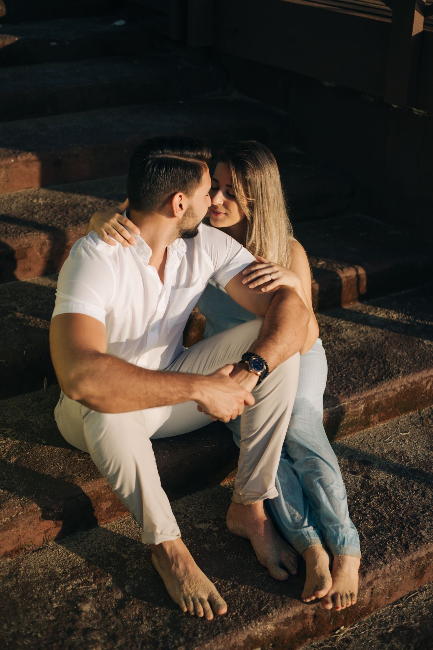 Loving couple embraces while sitting on outdoor brick steps in casual summer attire.