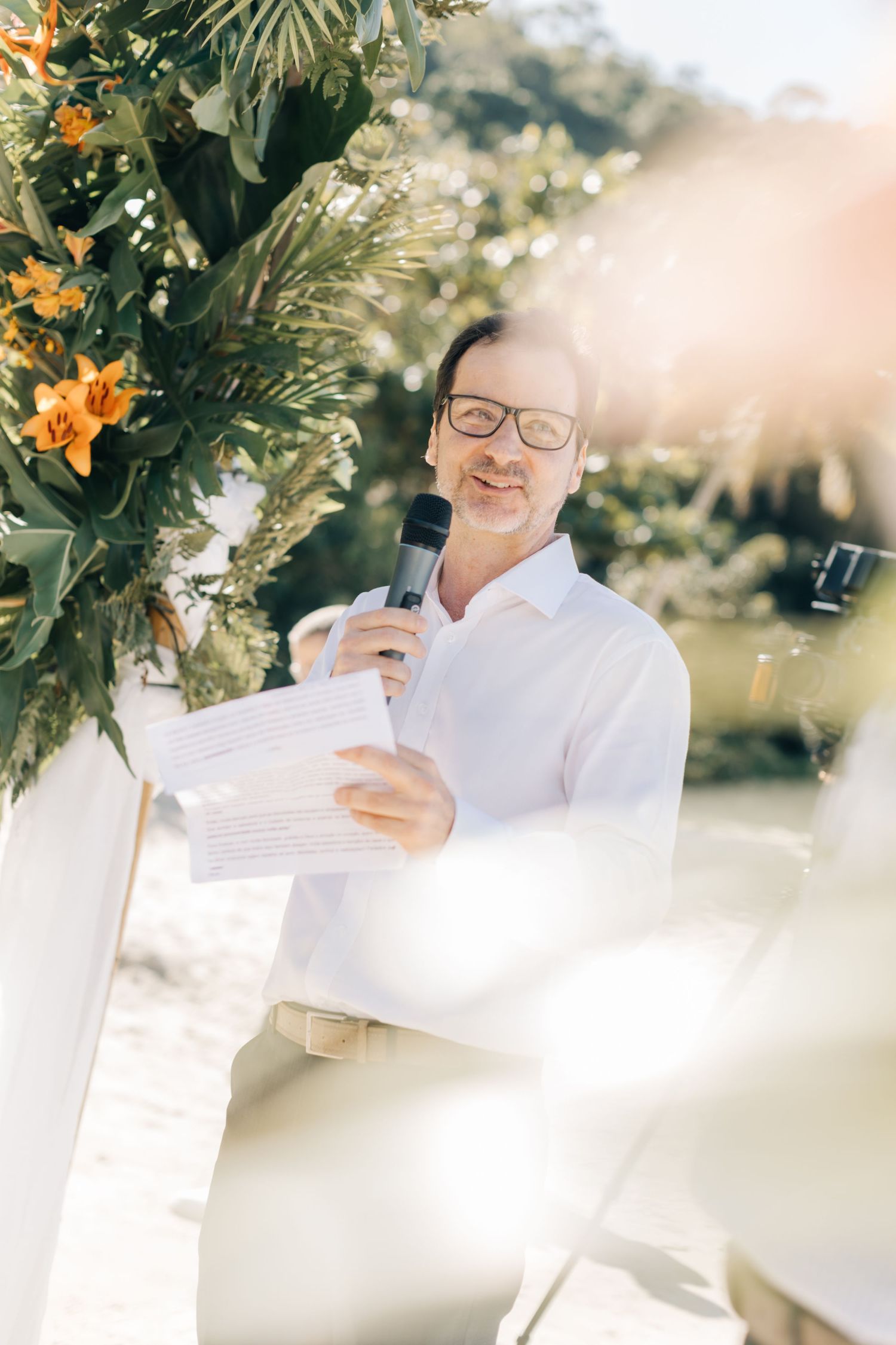 A wedding officiant in a white shirt delivers a speech outdoors with floral arrangements in the background.