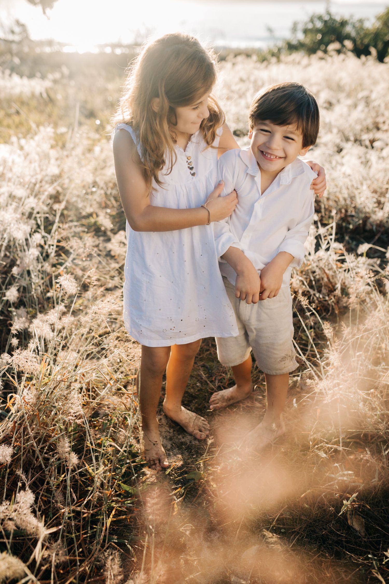 Children in white clothing play together in a sunlit field of golden grass and wildflowers.