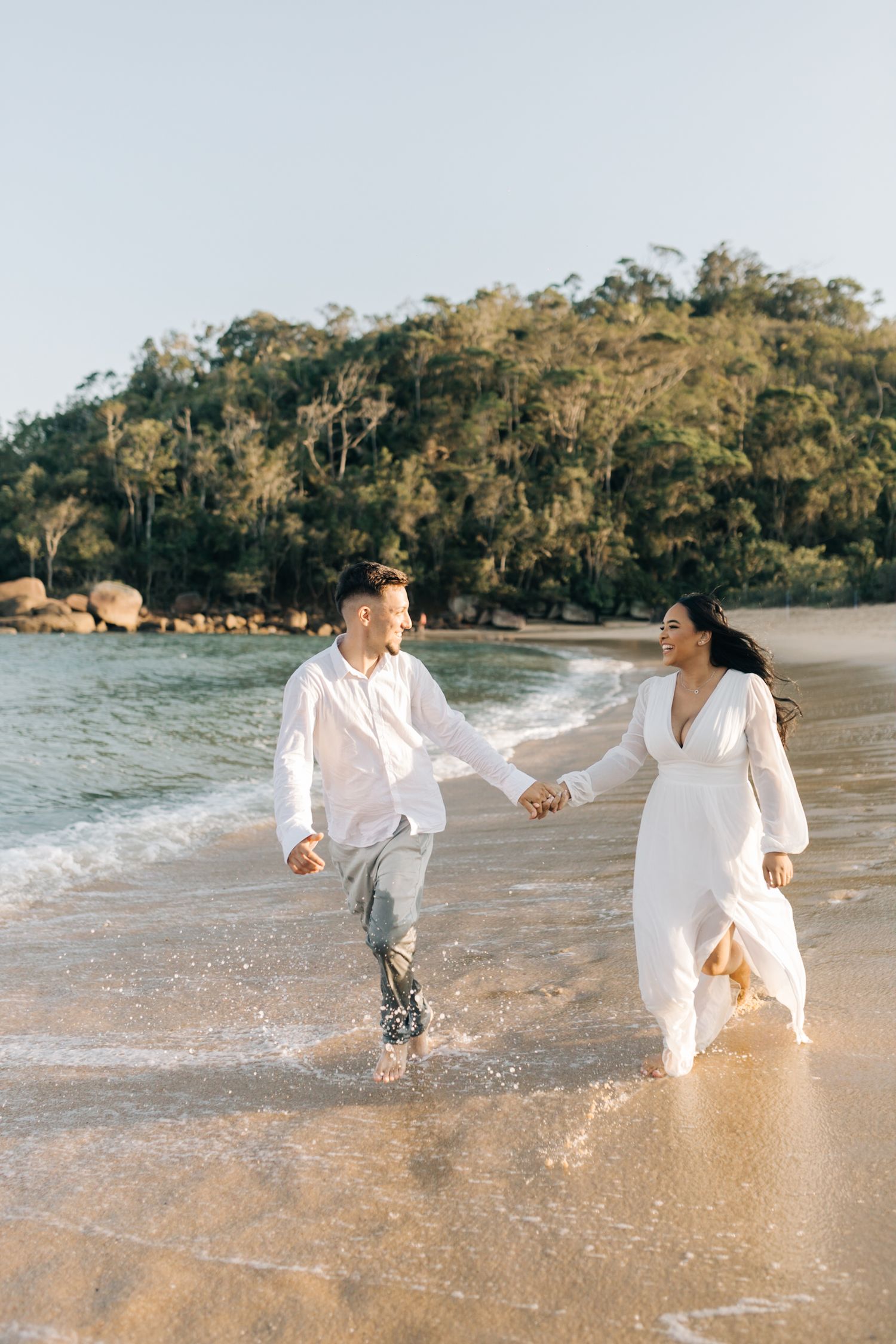 Joyful couple splashes through shallow ocean water in coordinated white clothing.