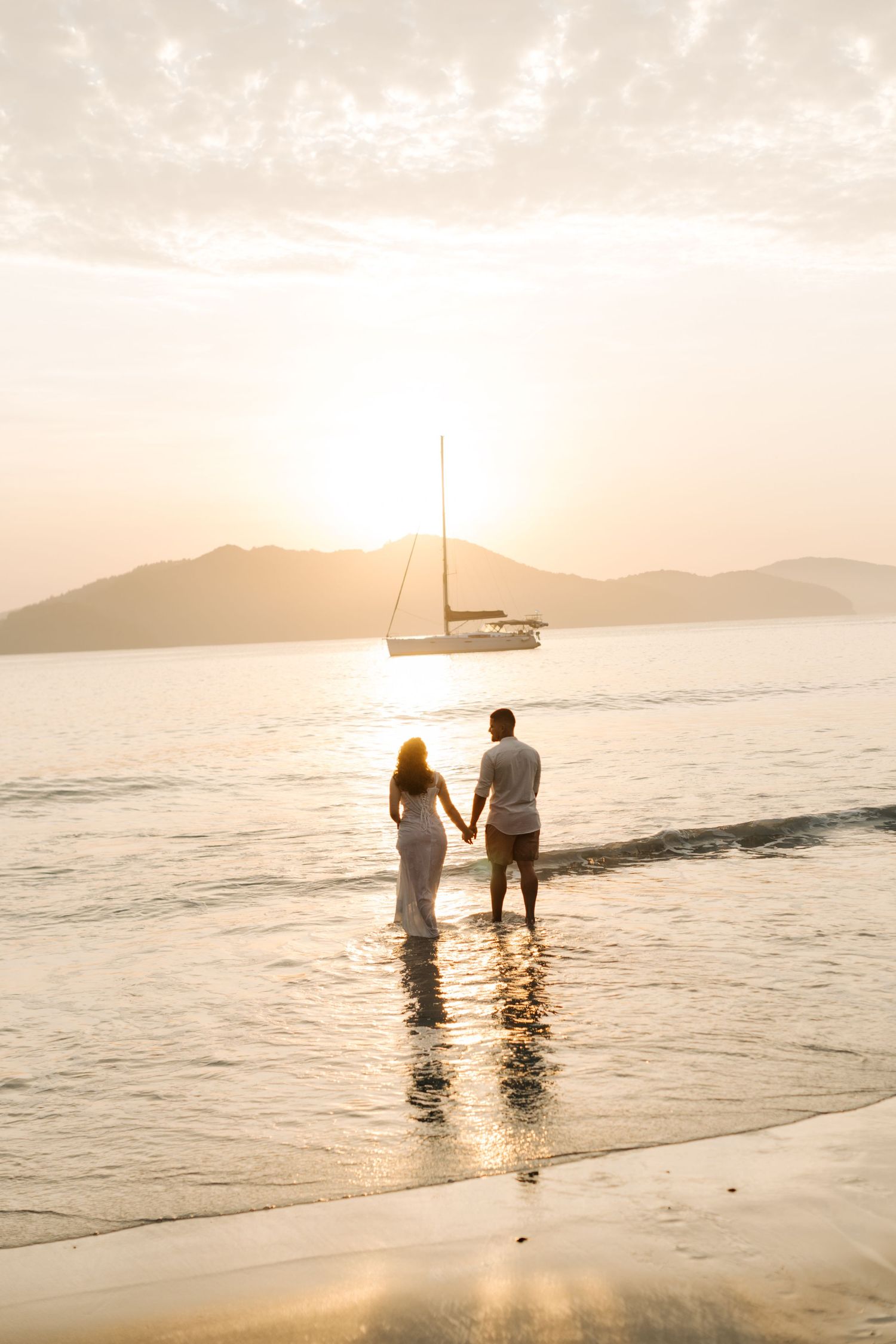Silhouetted figures wading in ocean water at sunset with sailboat and mountains in background.