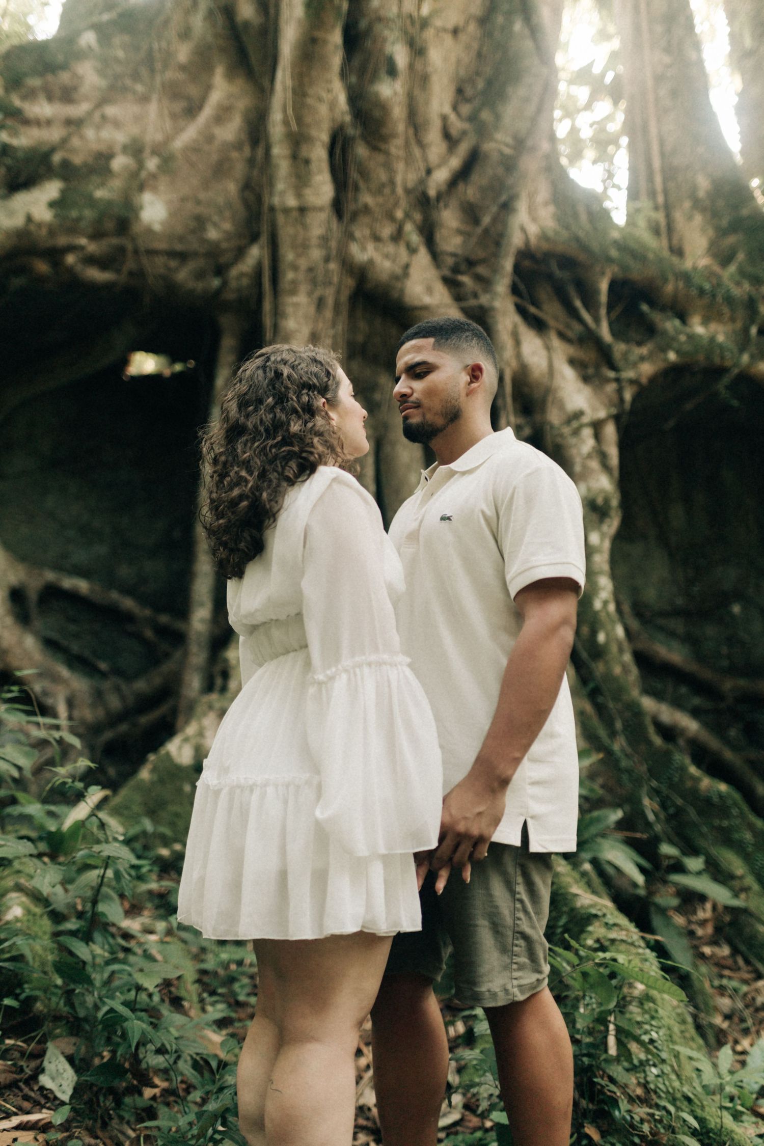Couple dressed in white clothing embrace tenderly among tree roots in a forest setting.