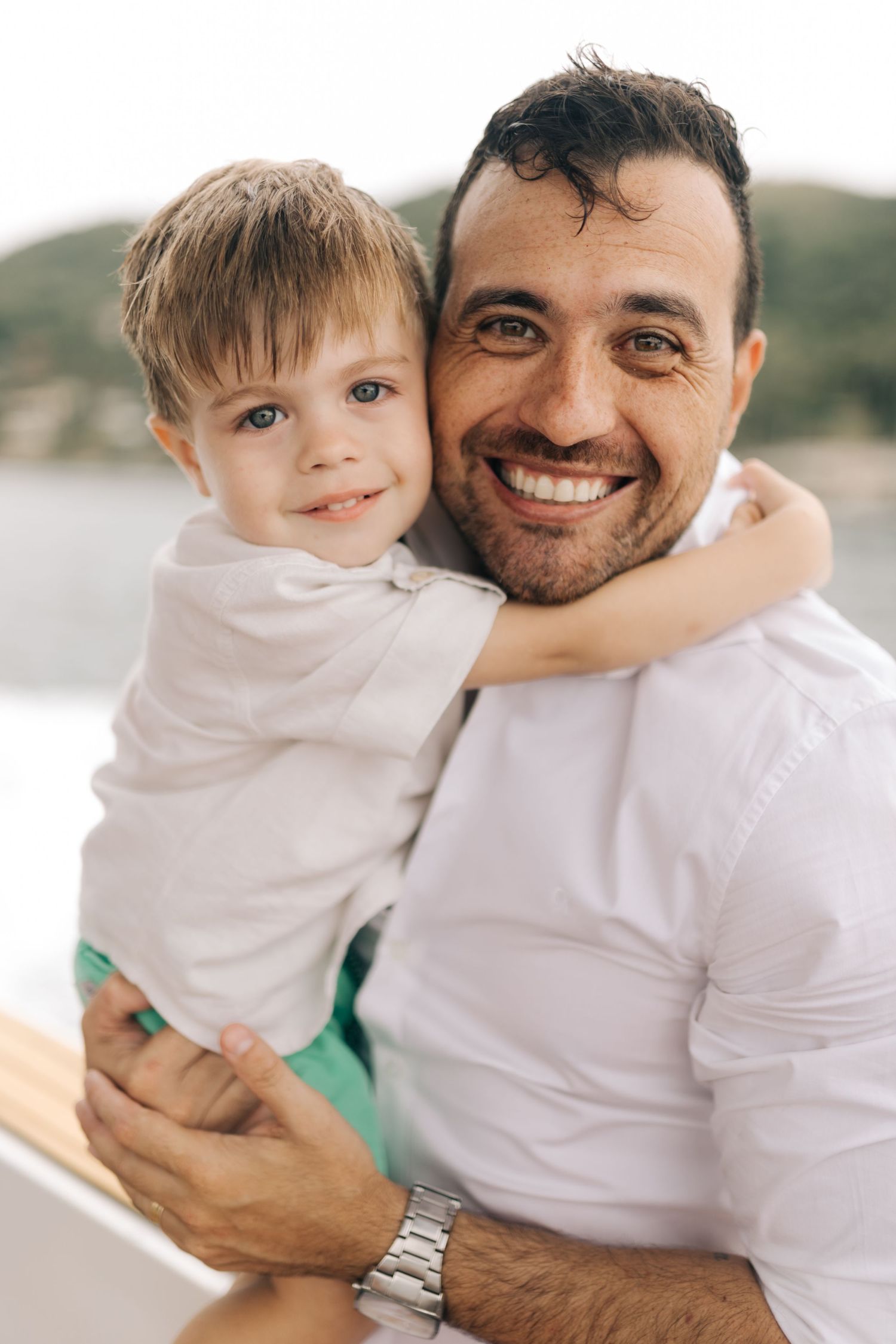 Parent and child in white shirts smiling together on boat with coastal background.