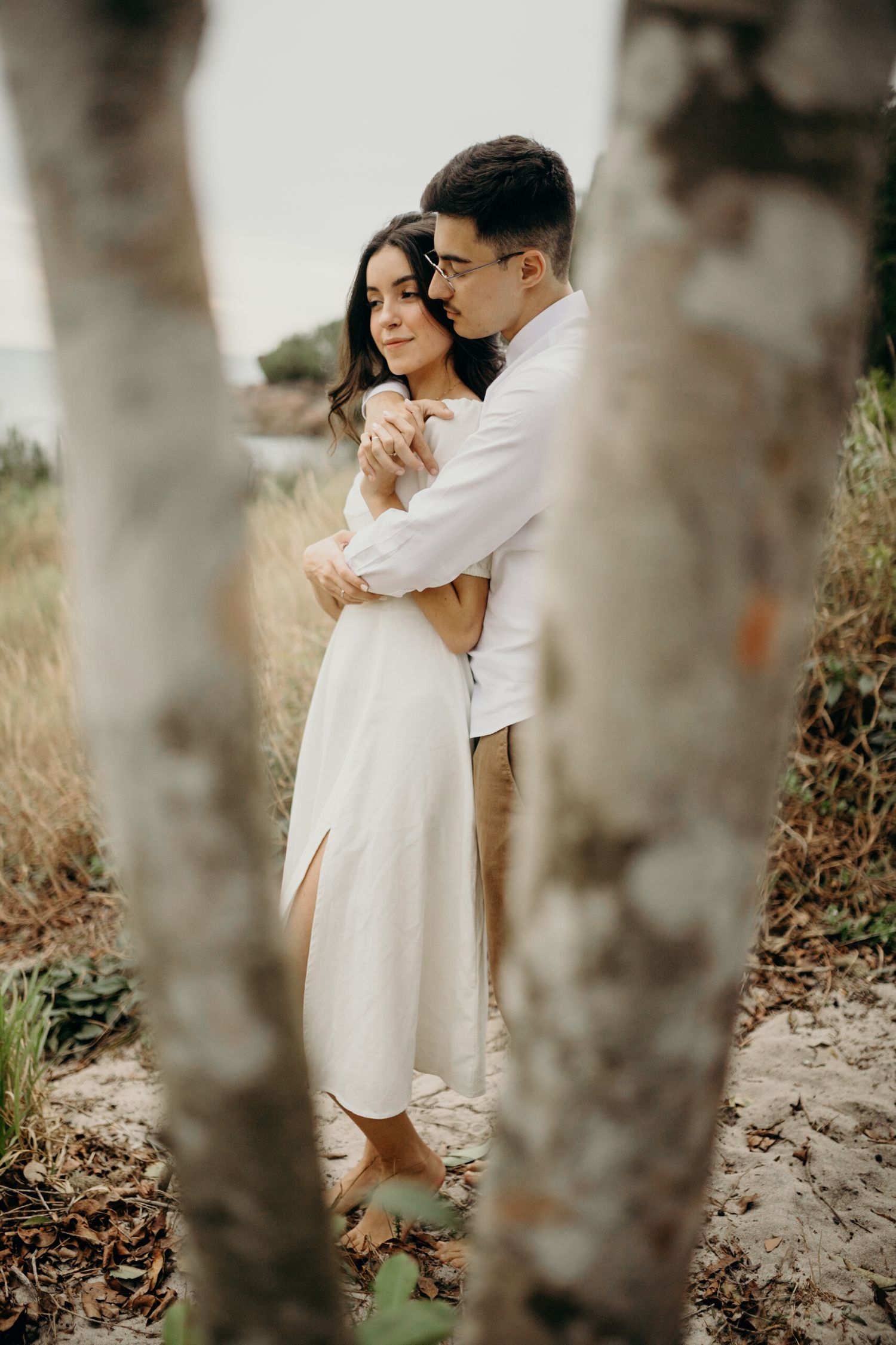 Romantic couple in white embraces tenderly in a wooded outdoor setting.