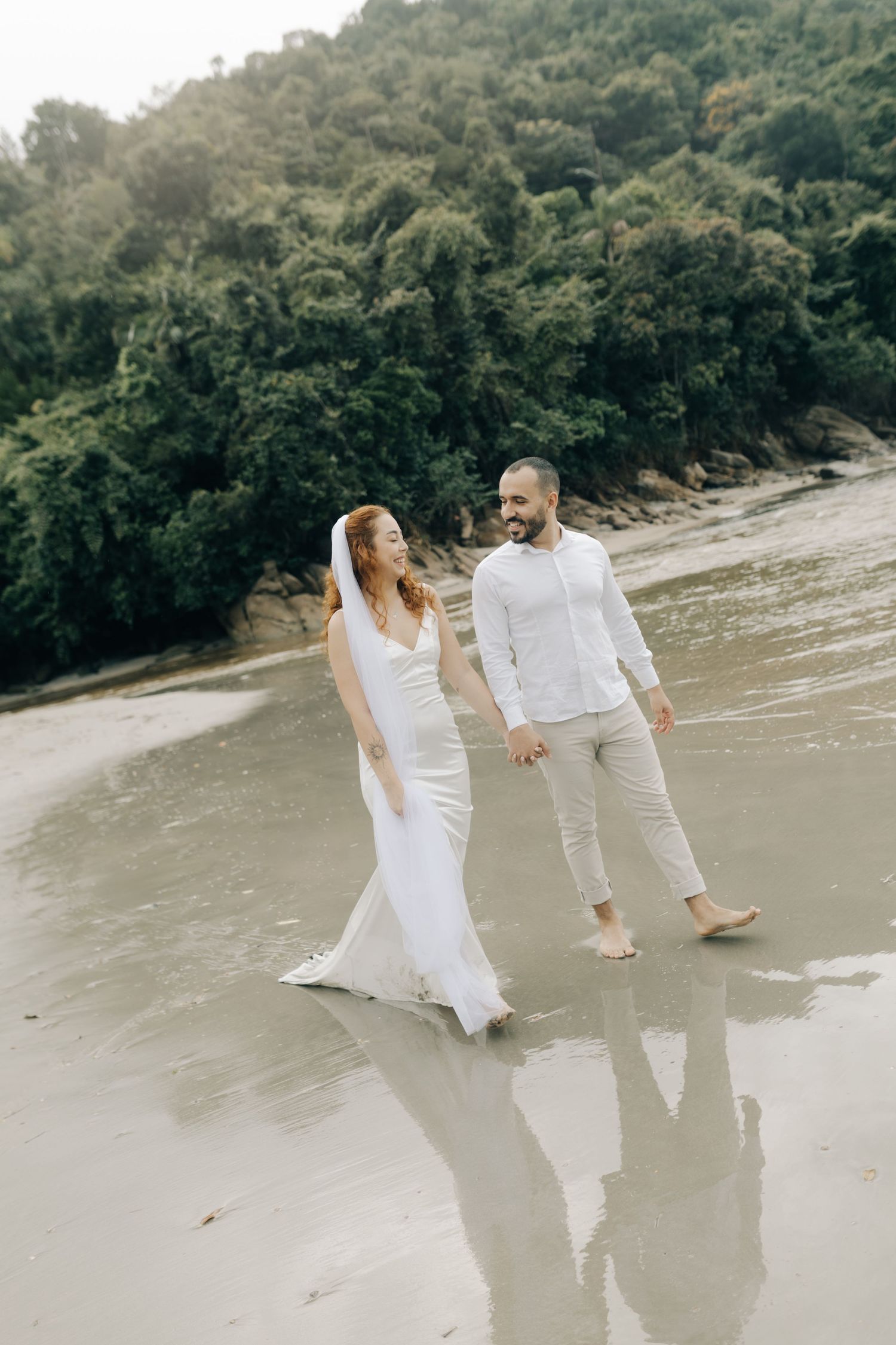 Newlyweds walking barefoot along scenic beach shore in white attire.