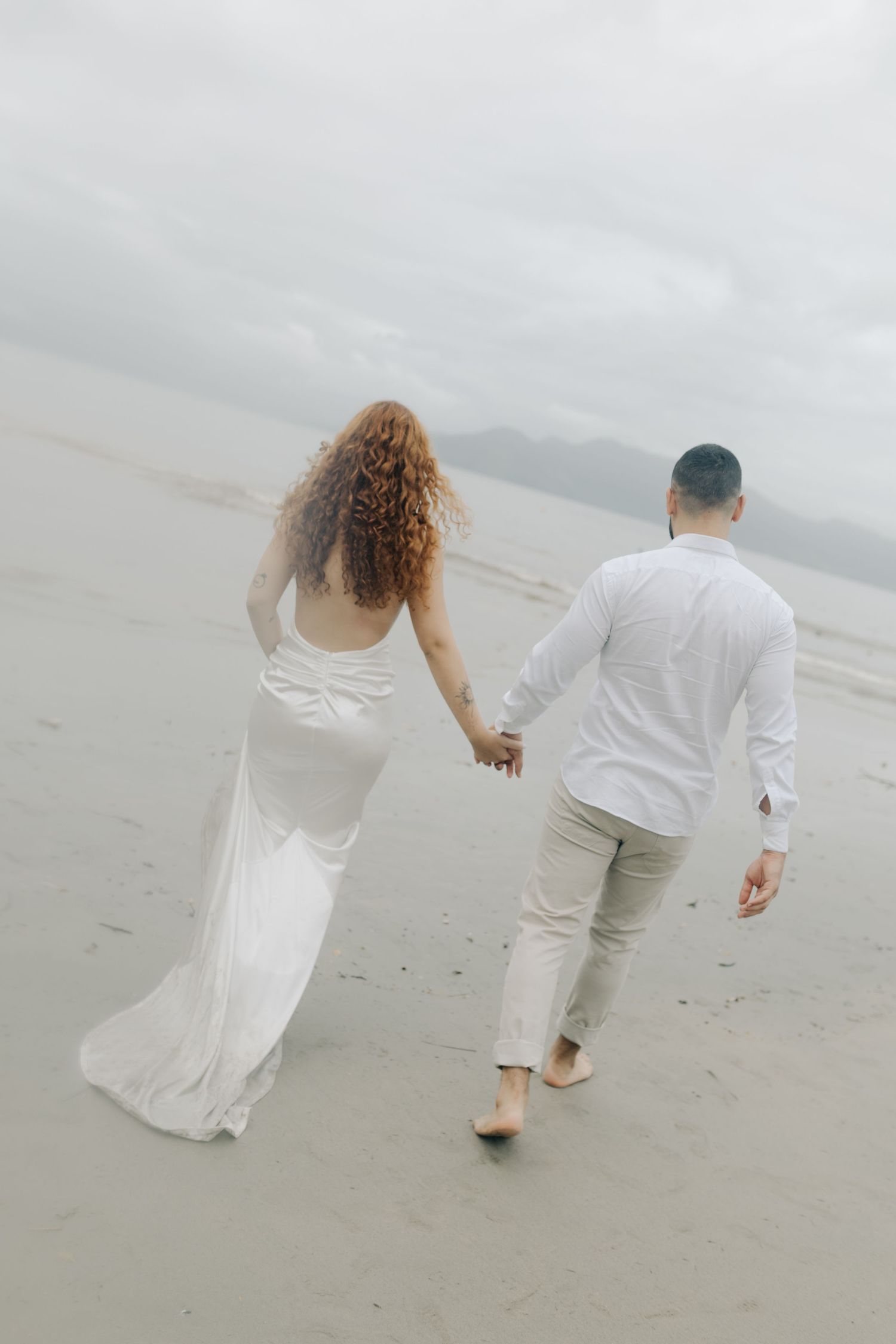 A couple in white clothing walks hand in hand along a foggy beach.