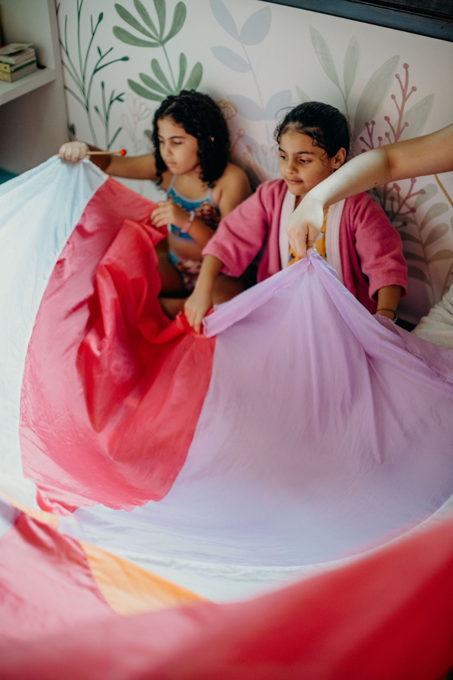 Two people relax on a bed with colorful sheets against a wall with floral designs.