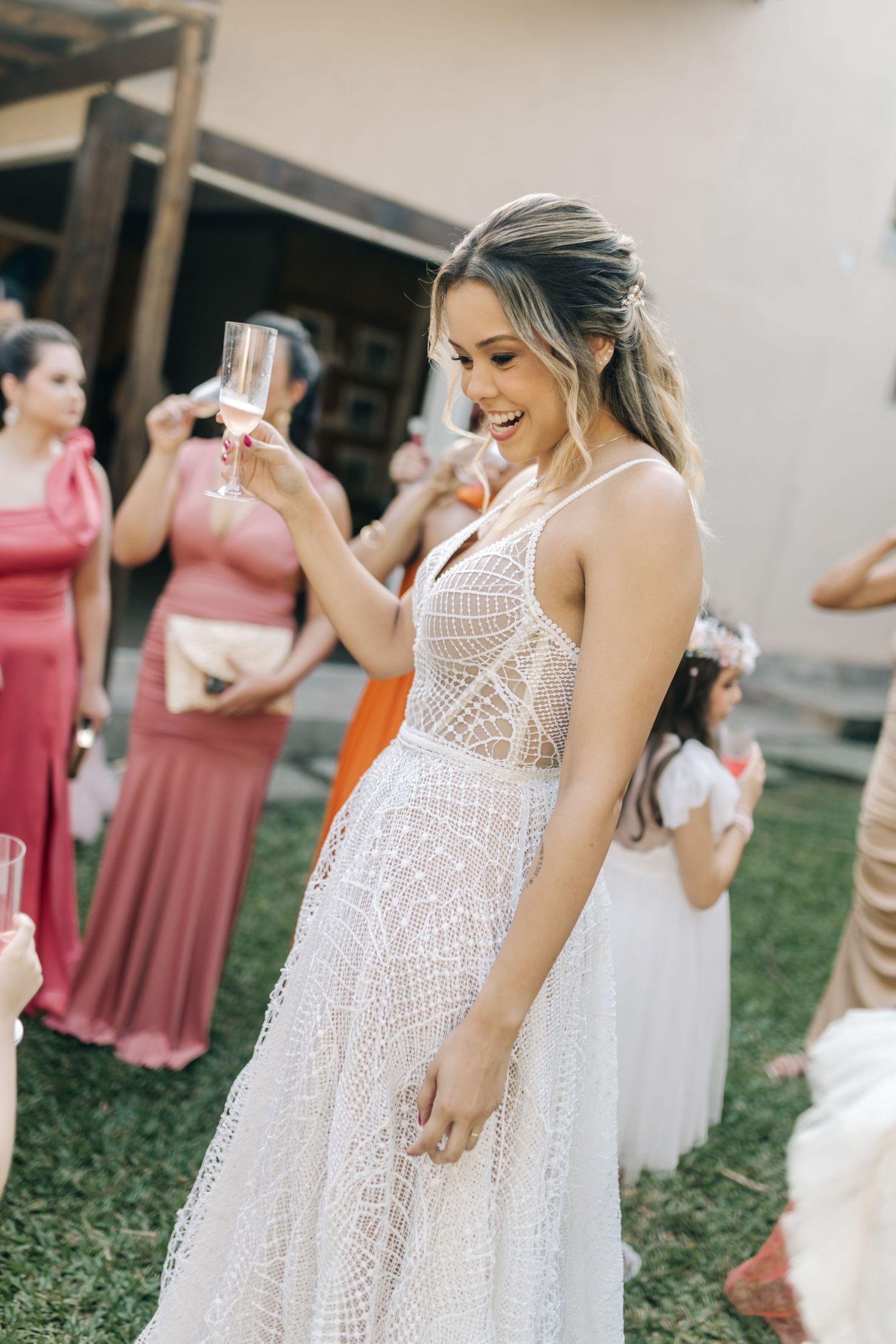 A person in a detailed white lace wedding gown smiles while holding a drink at an outdoor reception.