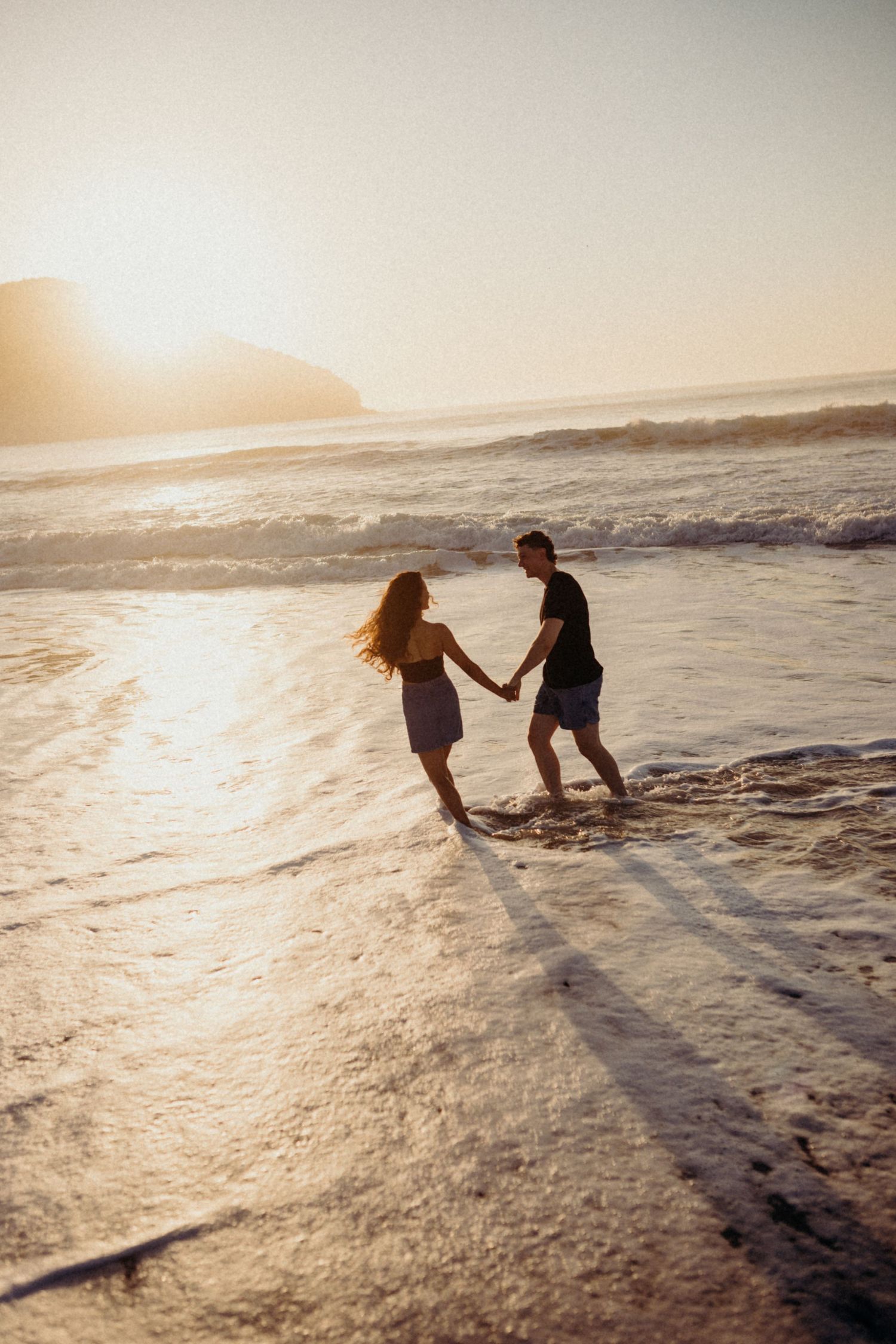 Couple holds hands while playing in shallow ocean waves during golden hour sunset.