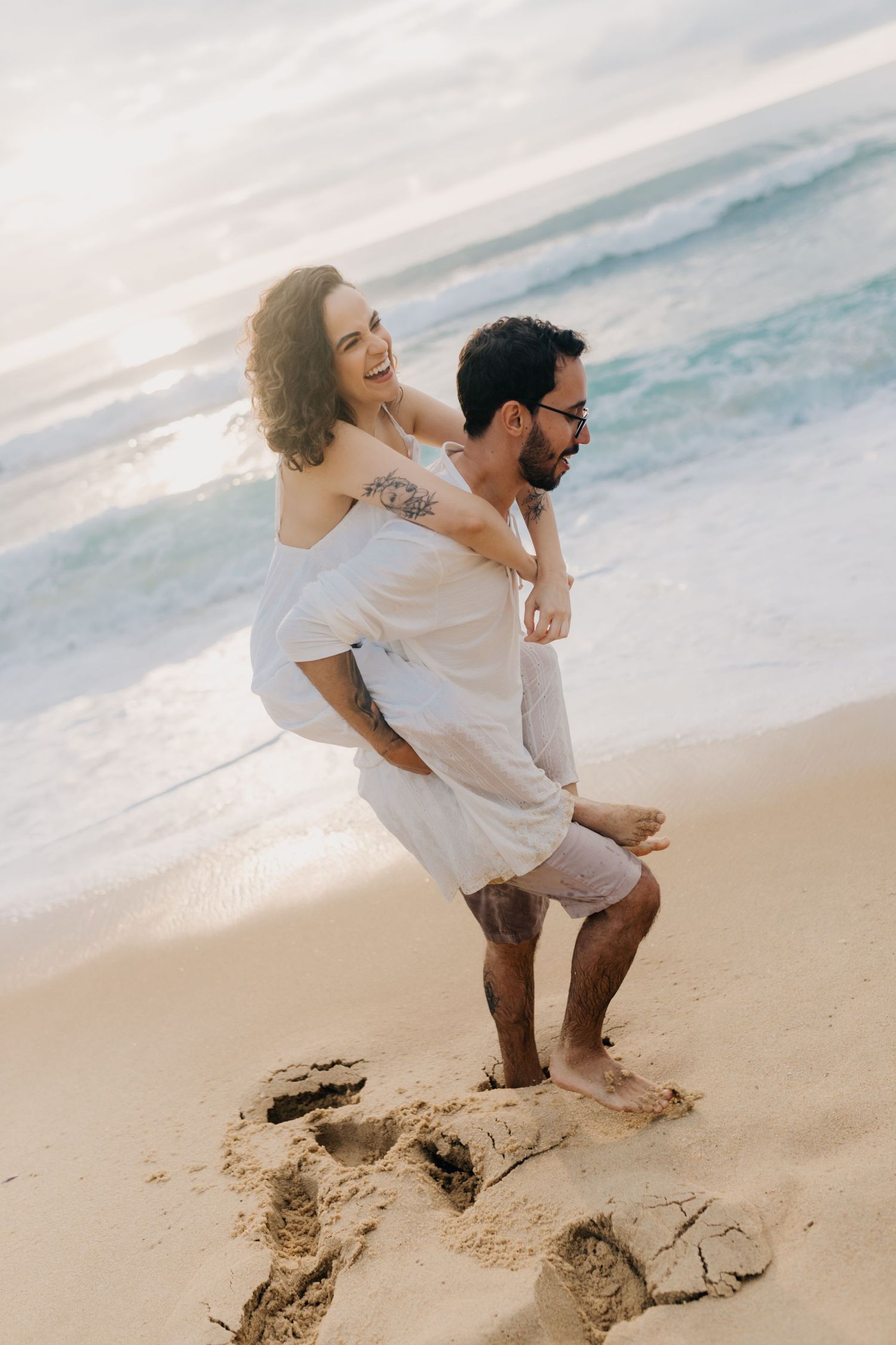 A couple shares a joyful piggyback ride moment on a sandy beach during sunset.