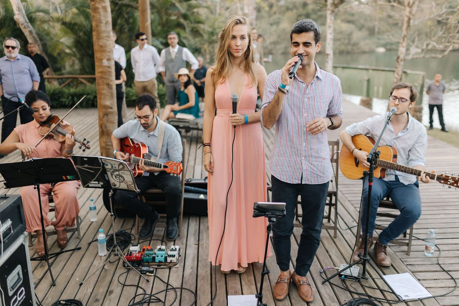 Musicians perform live music at an outdoor wedding venue with acoustic instruments and microphones.
