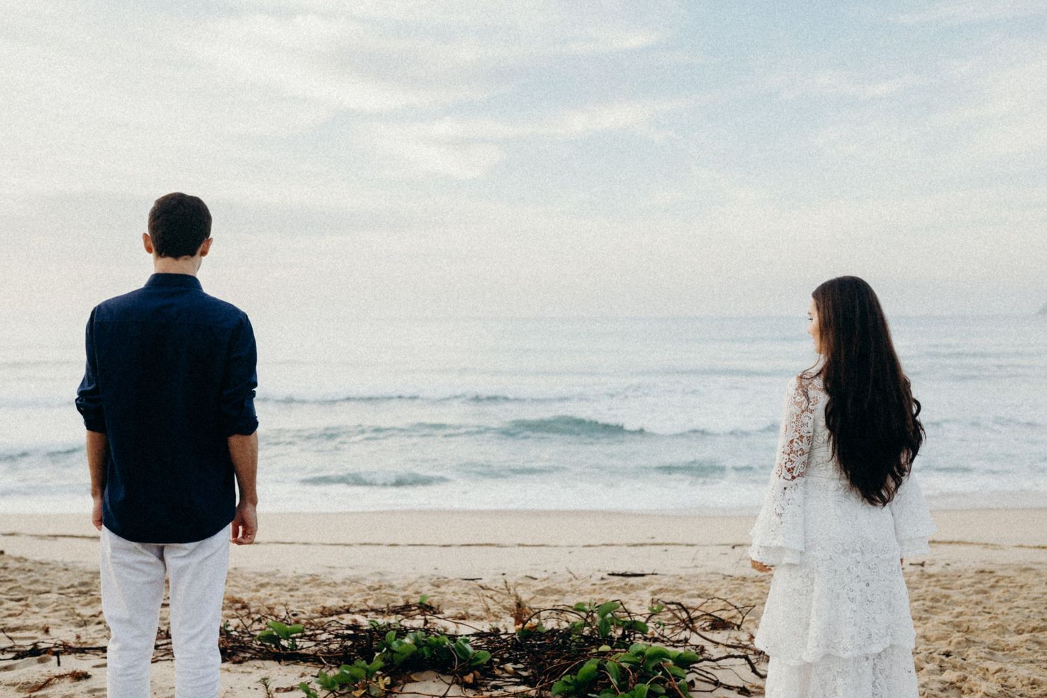 A couple in coordinated navy and white outfits stands apart while gazing at the ocean waves from a sandy beach.