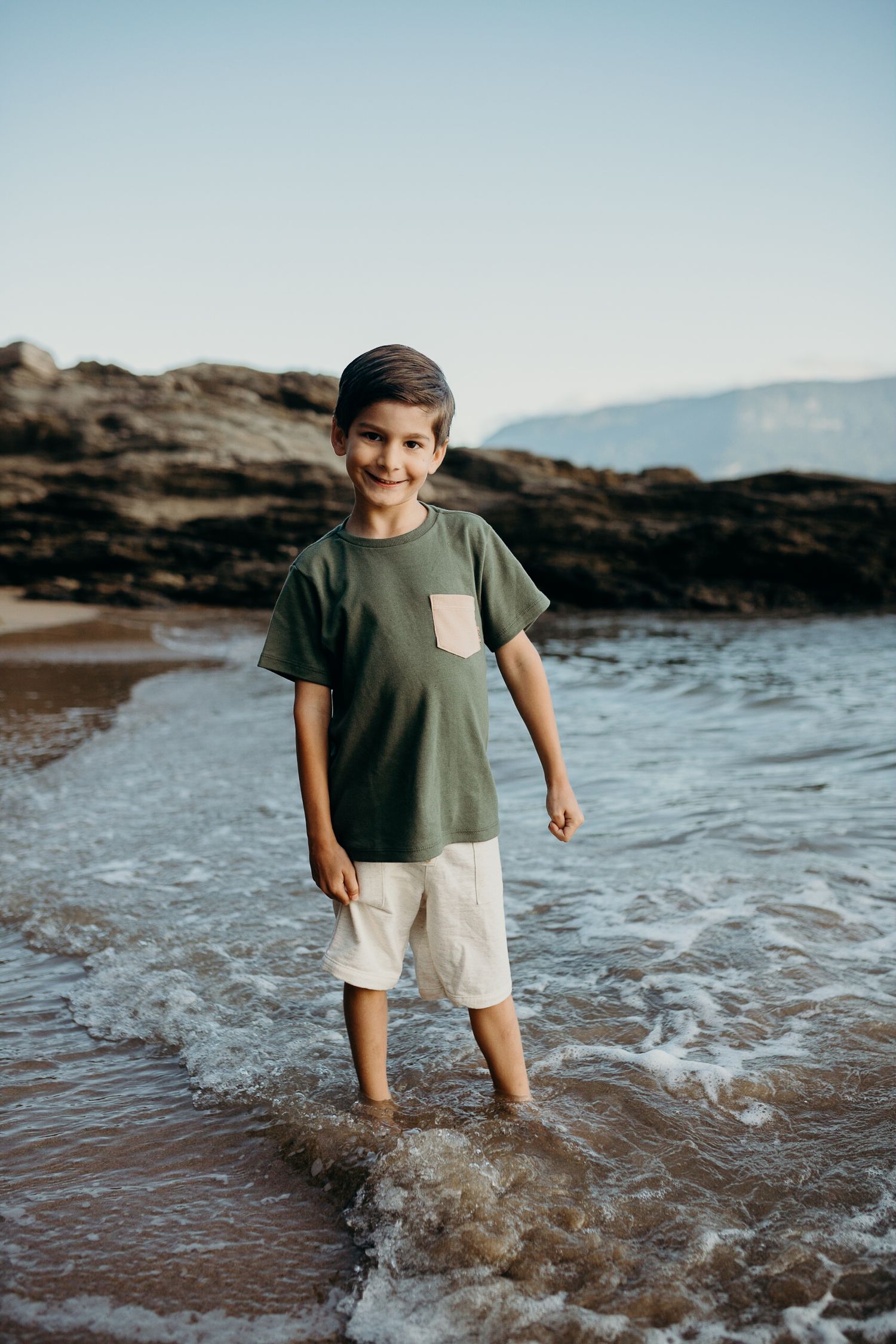 Young child standing in shallow ocean water at a rocky beach with mountains in background.