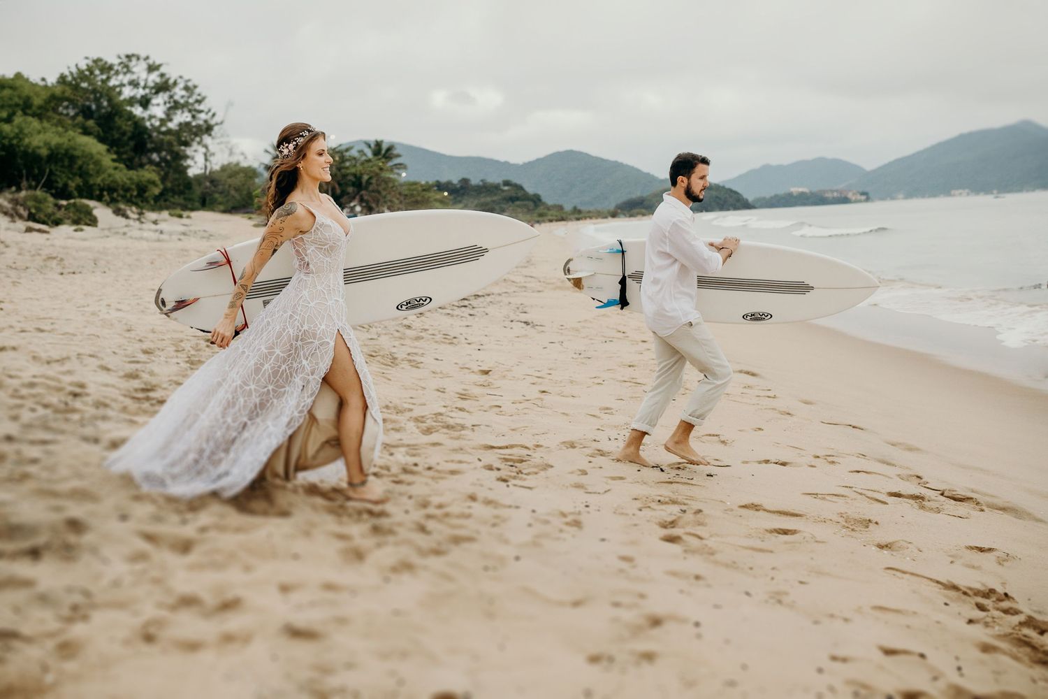 Couple in white outfits running playfully along a scenic beach with mountains in the background.