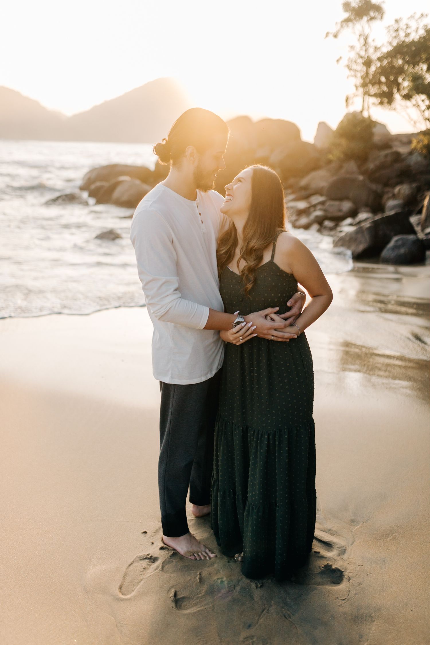 Romantic beachside portrait of couple embracing against backdrop of golden sunset and coastal rocks.