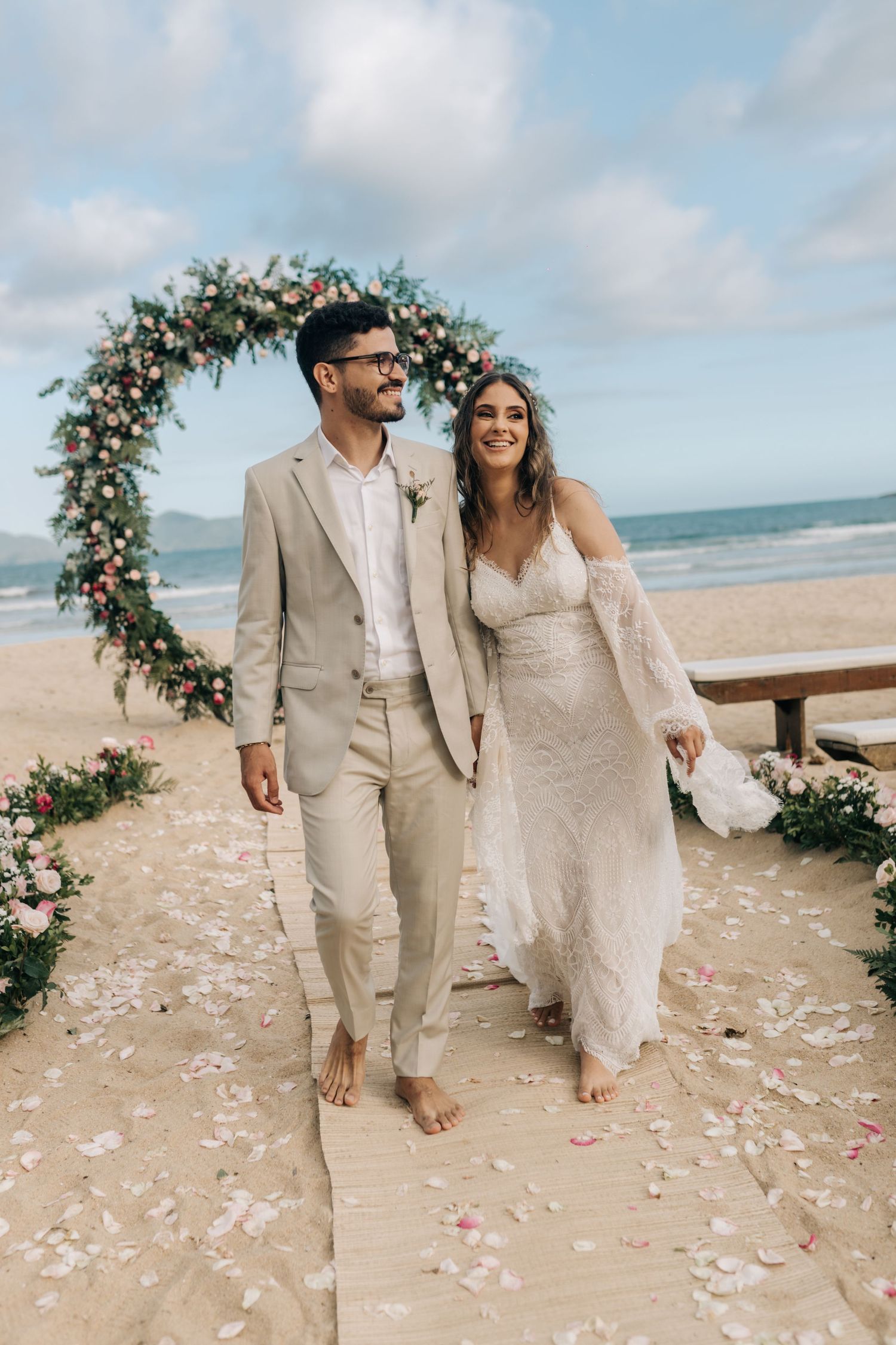 Barefoot newlyweds walk under a floral arch on a beach boardwalk with rose petals scattered.