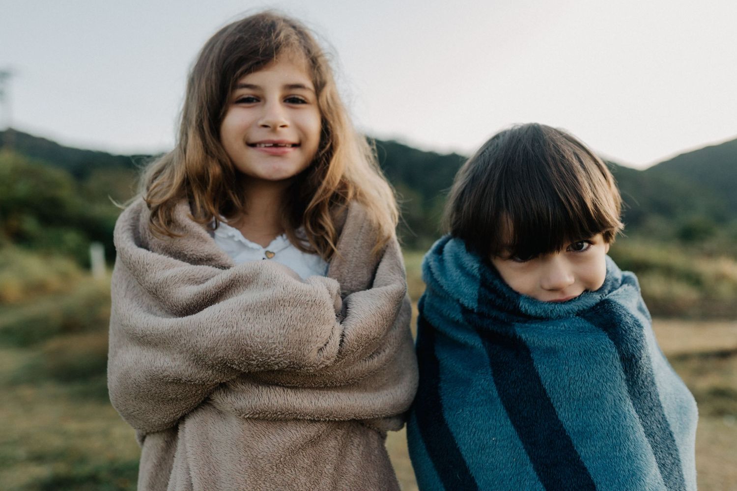 Two kids wrapped in blankets smiling together outdoors.