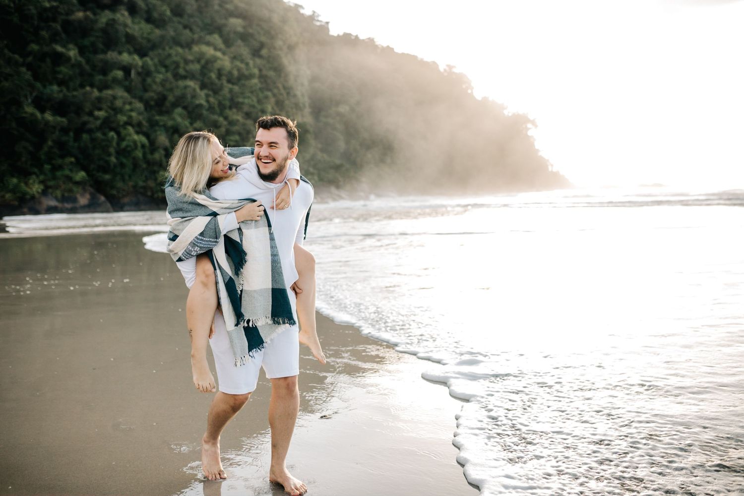 Couple walking along ocean shore during golden hour with dramatic cliffside backdrop.