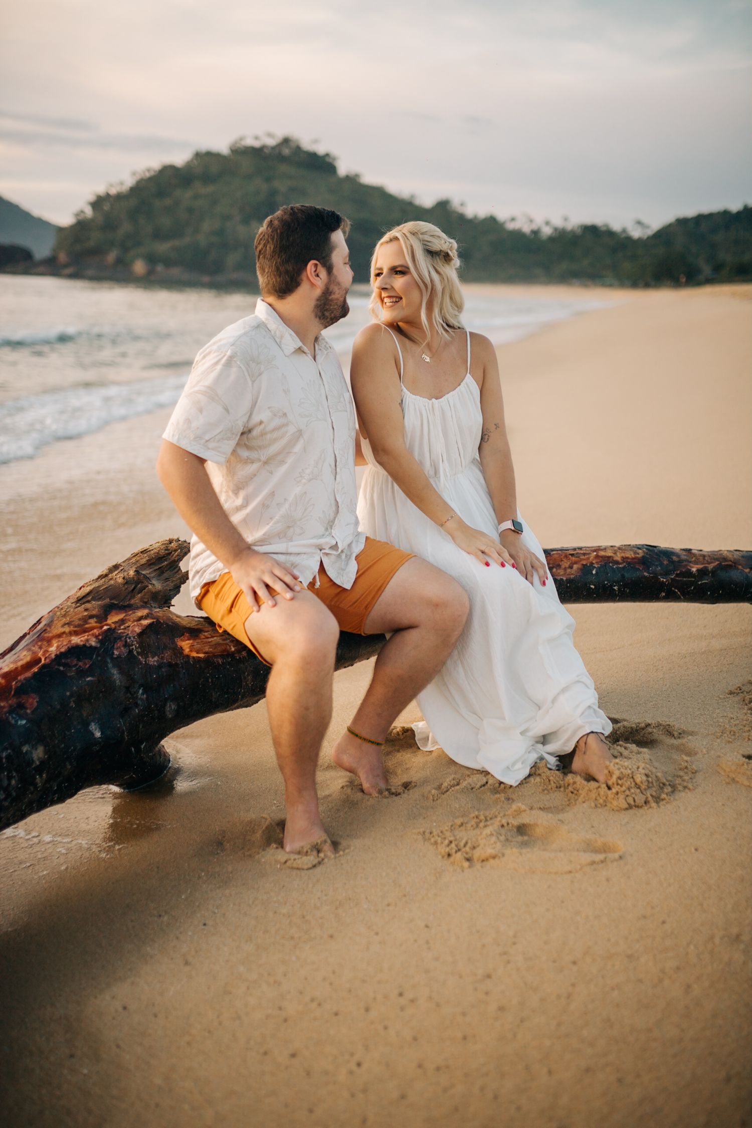 A romantic couple sits on driftwood at sunset on a beach with mountains in the background.