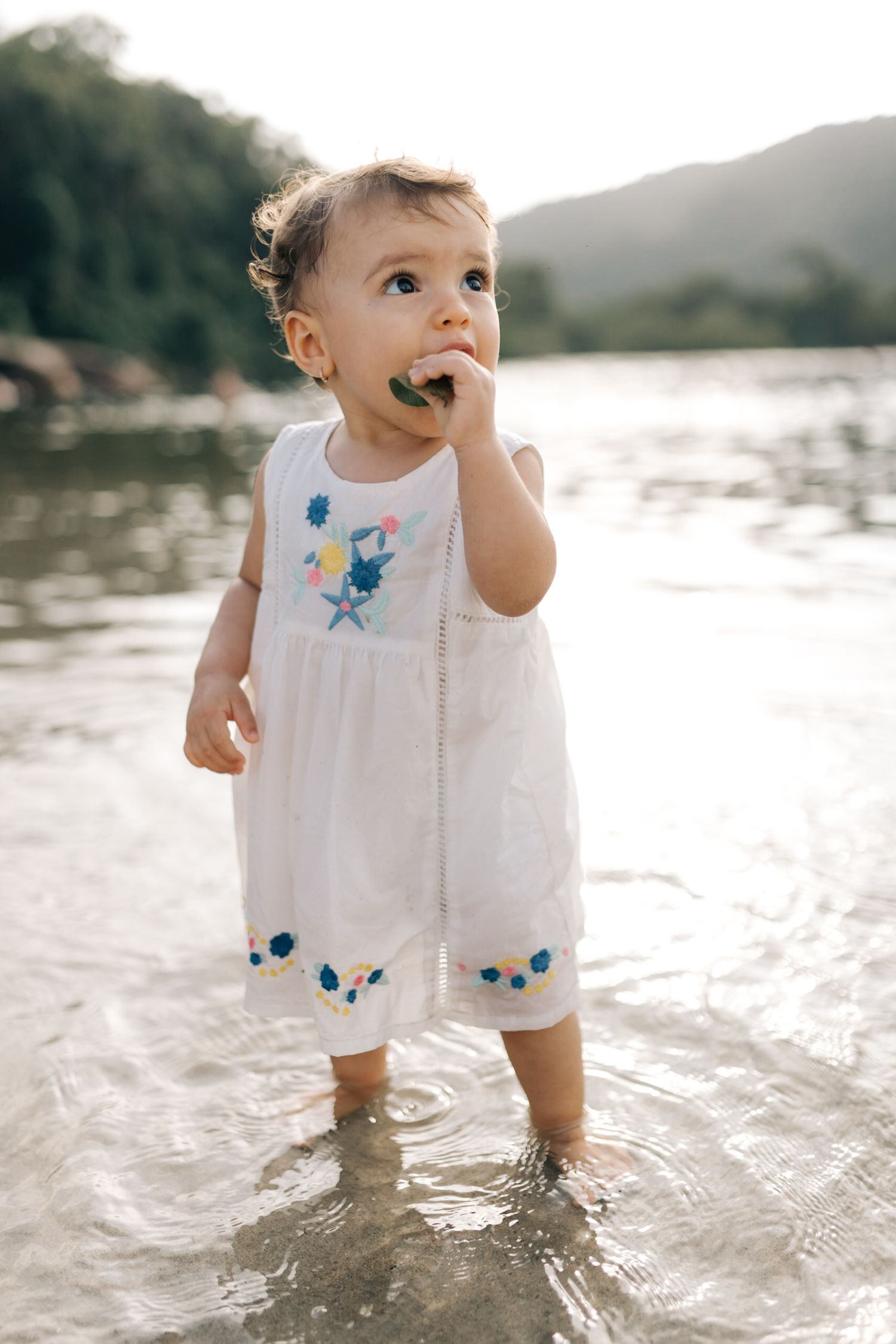 A toddler in a white dress with blue details splashes in shallow ocean water during sunset.