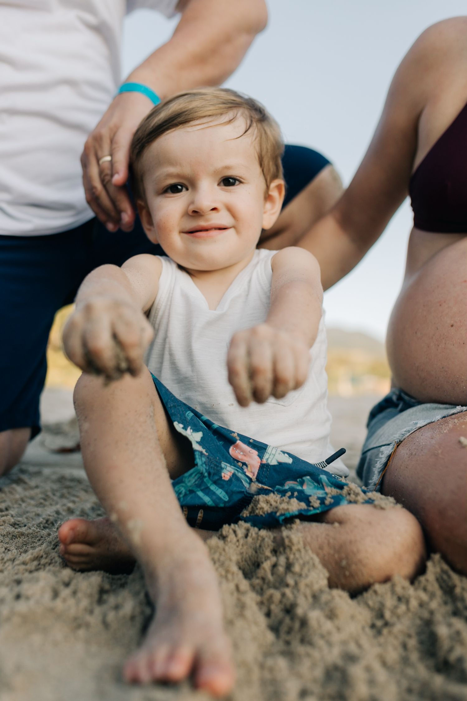 Young child plays in sand wearing blue shorts and white tank top during beach outing.