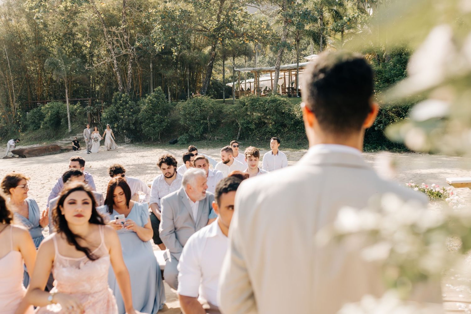 Wedding guests gather outdoors on a sunny day with lush greenery in the background.