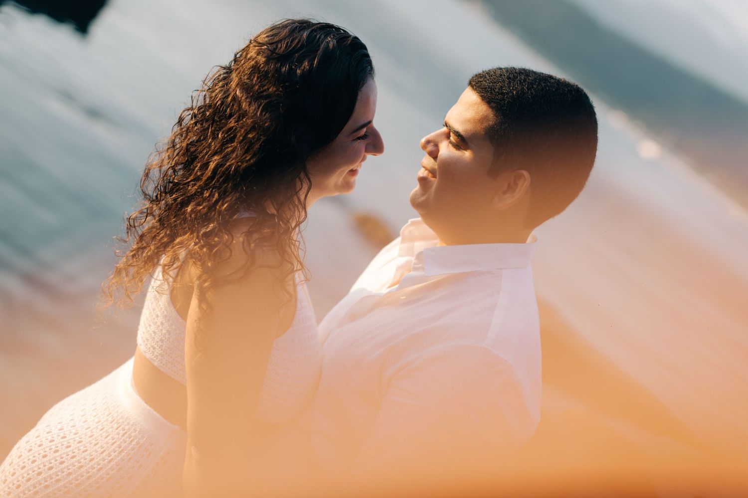 Romantic sunlit portrait of couple in white attire against ocean background.