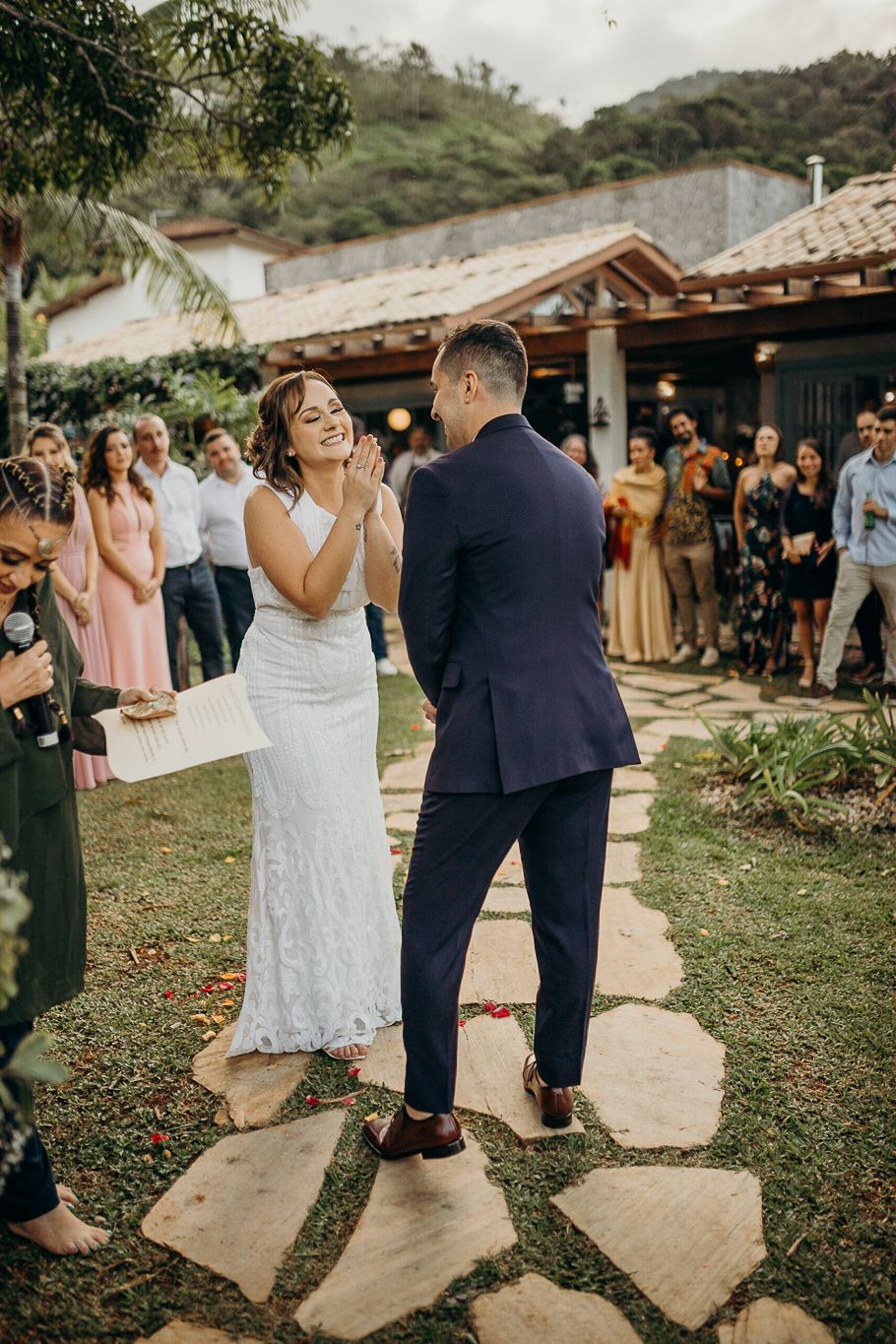 Guests observe a wedding ceremony on a stone pathway surrounded by lush greenery and rustic buildings.
