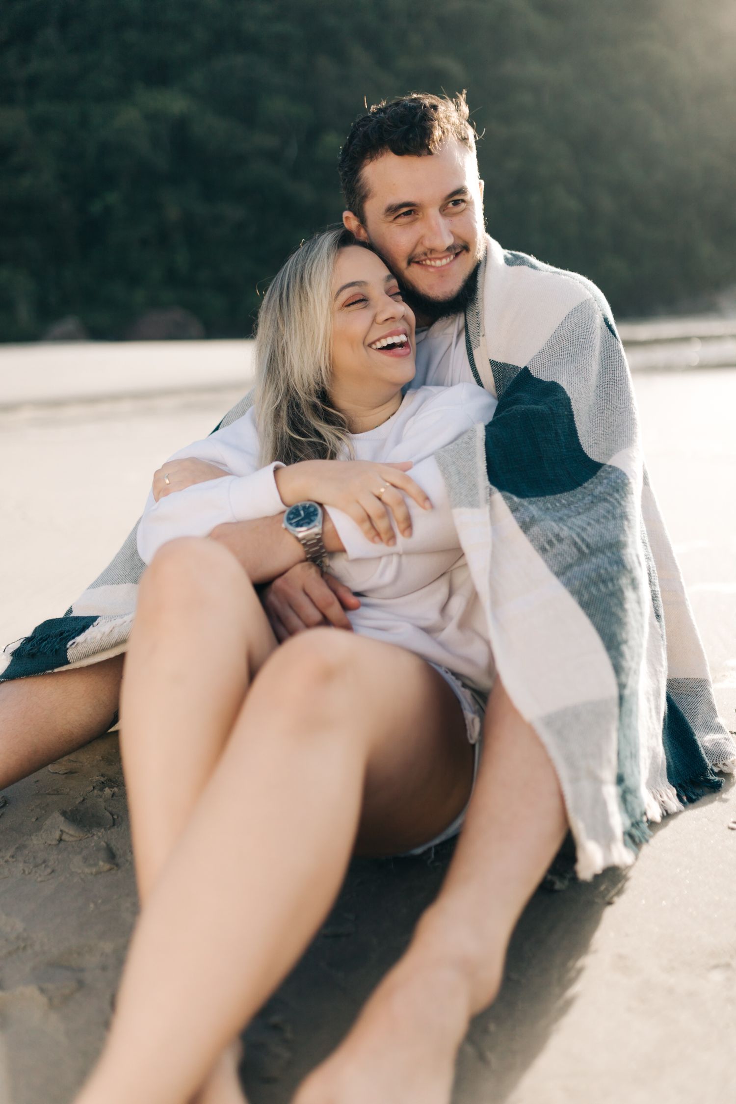 Joyful couple laughing together while relaxing on beach with cozy blanket.