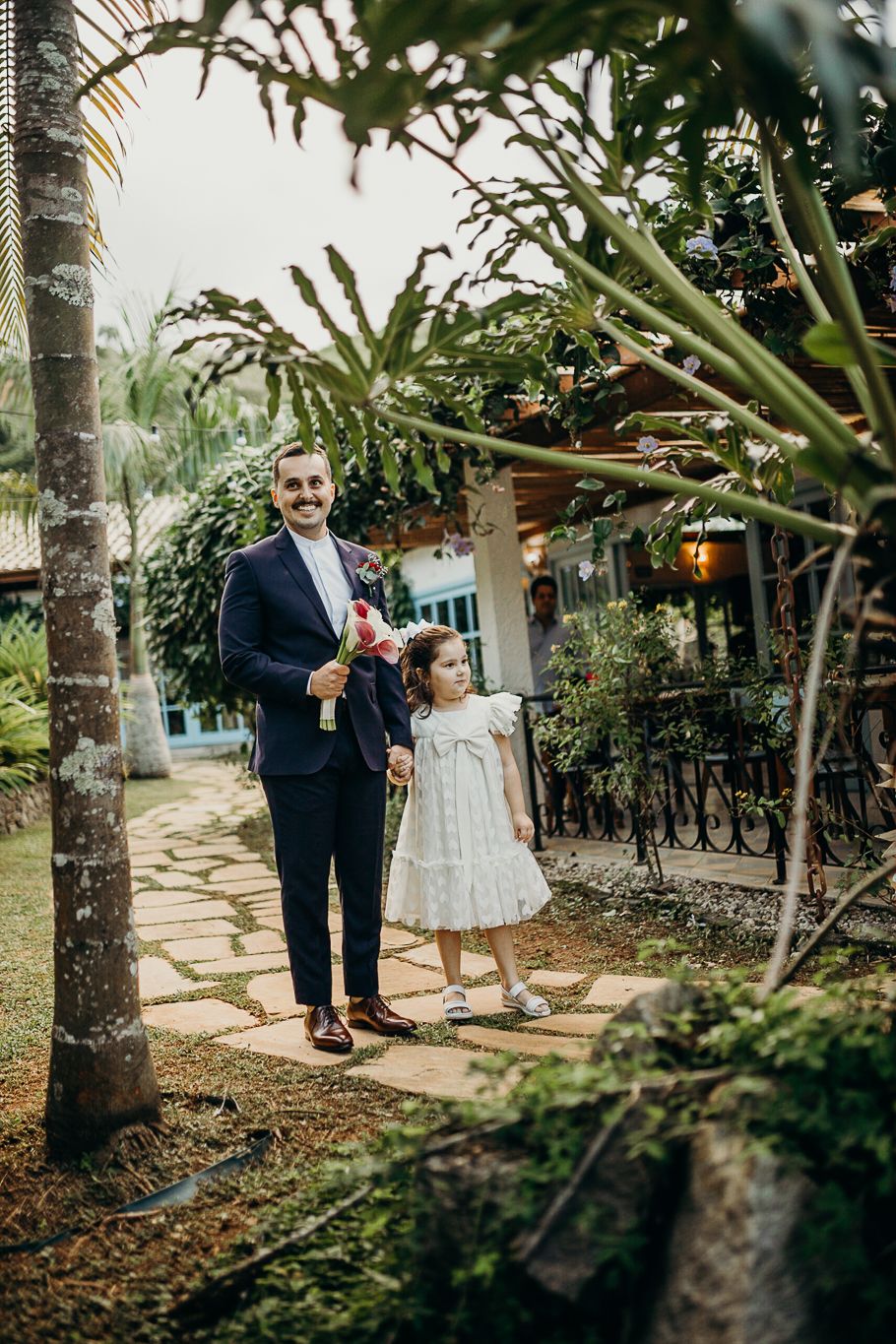 Formal dressed figures stand on garden pathway under tropical trees during outdoor event.
