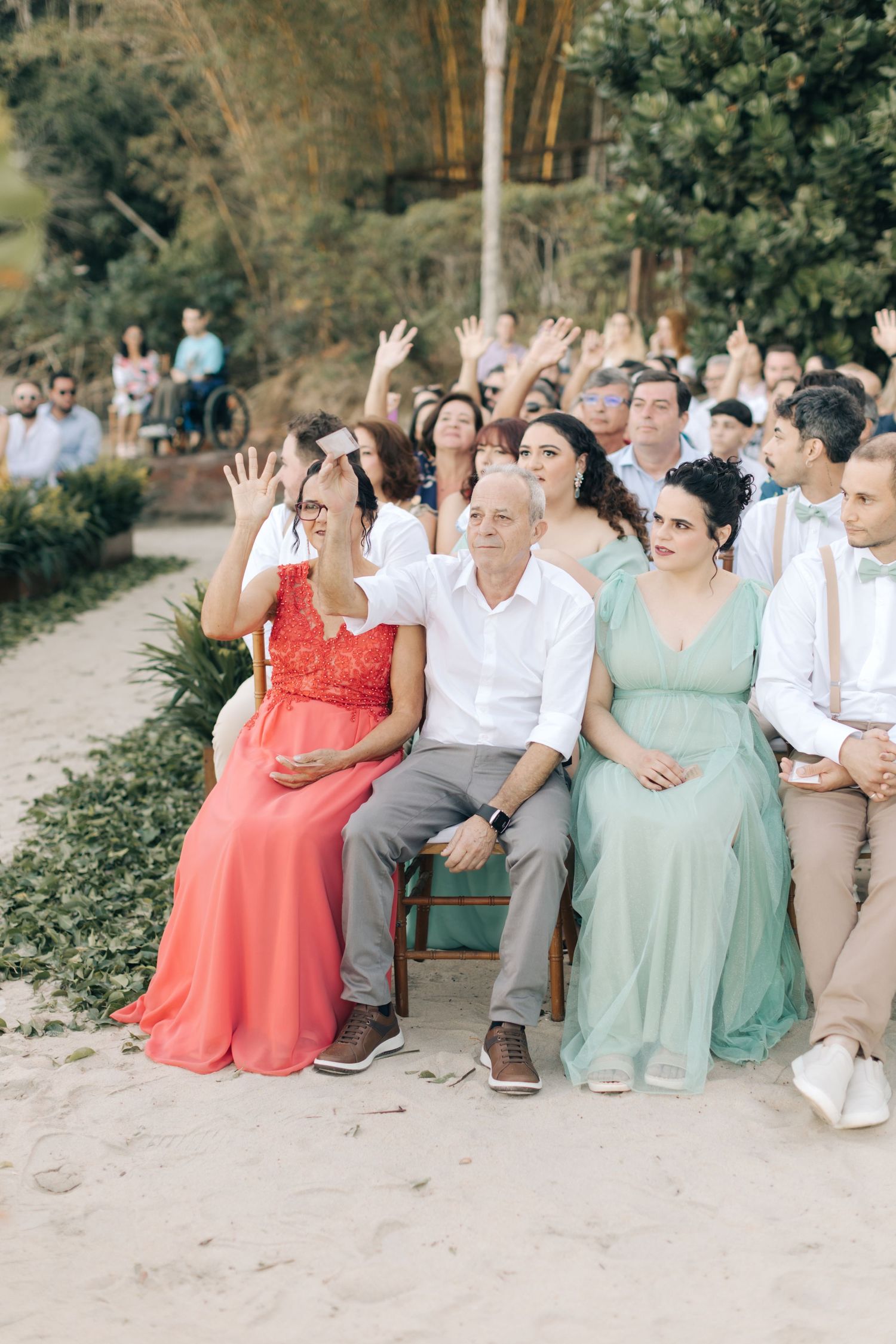 Wedding guests in colorful formal attire seated outdoors raise their hands during a celebration.