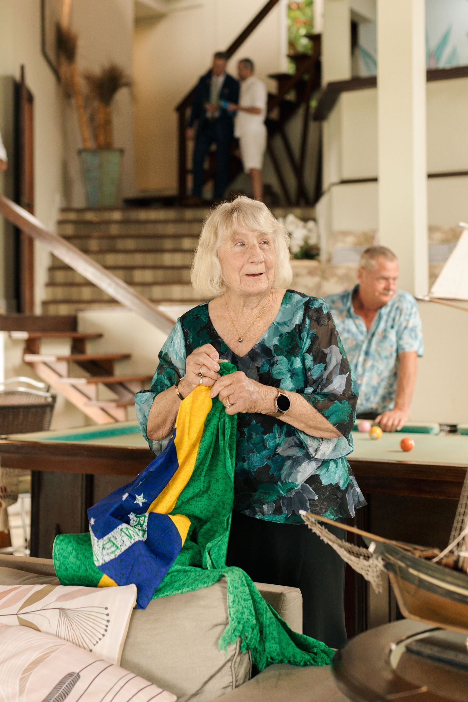A person in a teal top joyfully displays a Brazilian flag in a home setting.