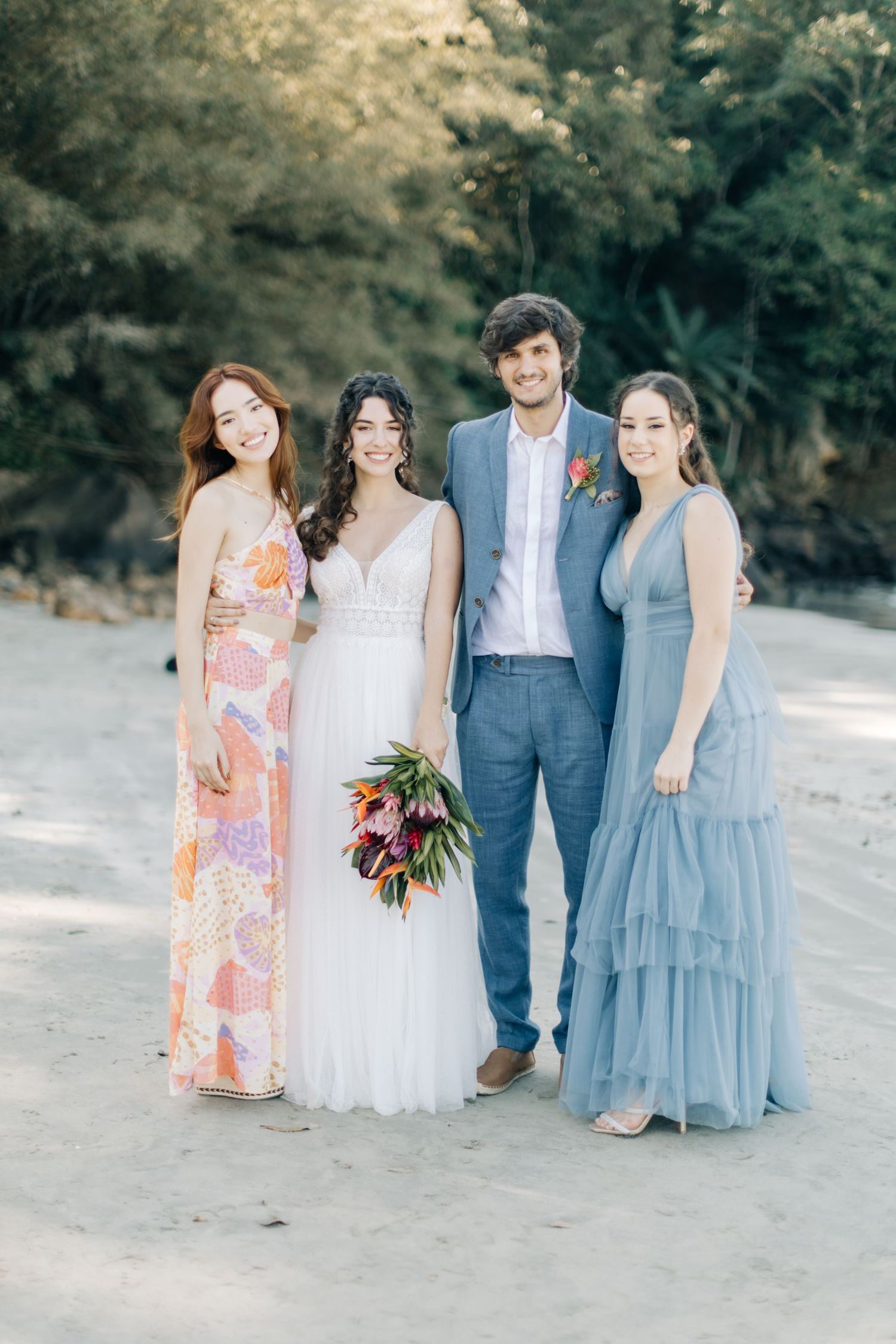 Wedding group in blue and floral dresses stand together on a beach with greenery behind them.