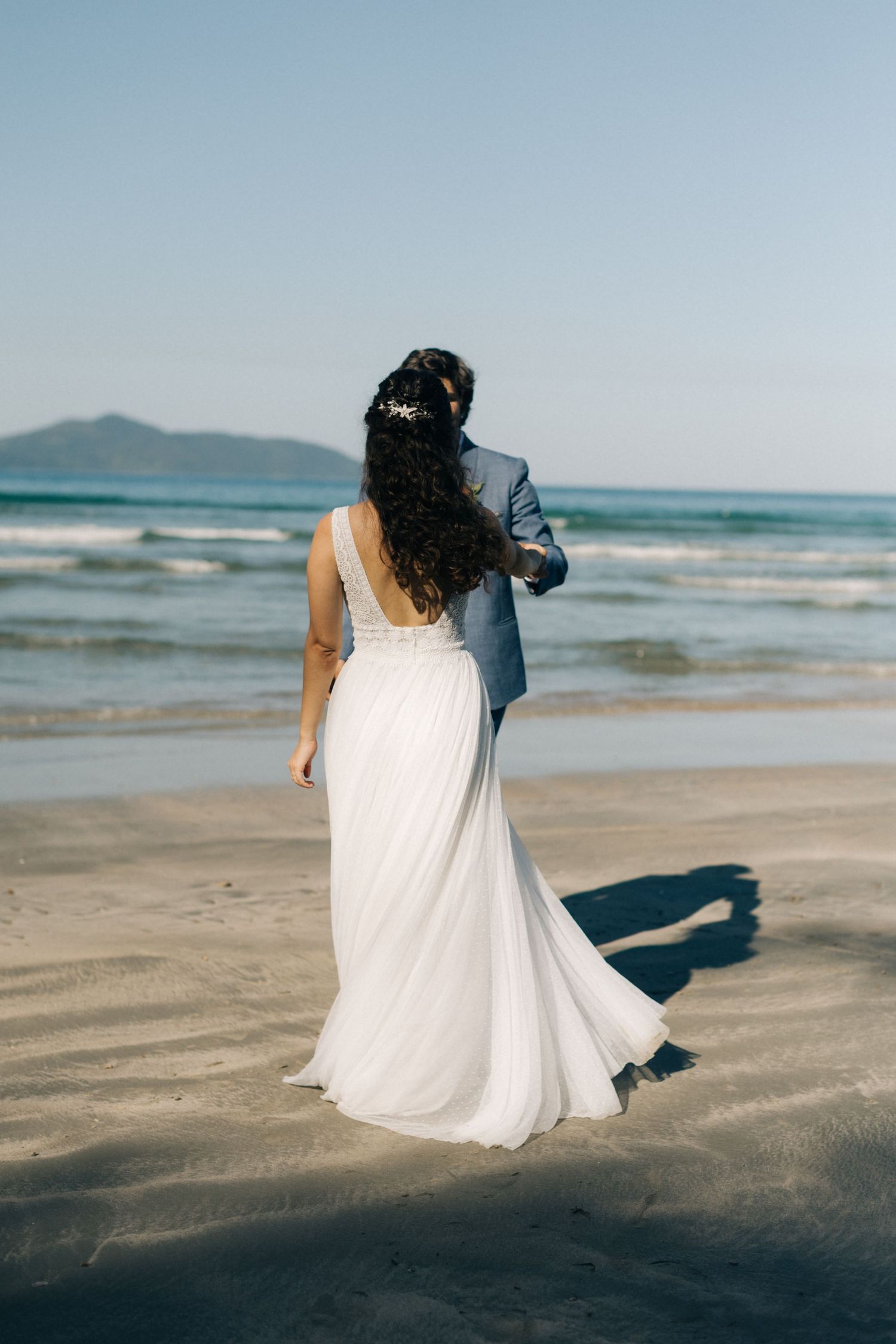 A flowing white wedding dress trails in the sand as newlyweds walk along a scenic beach.