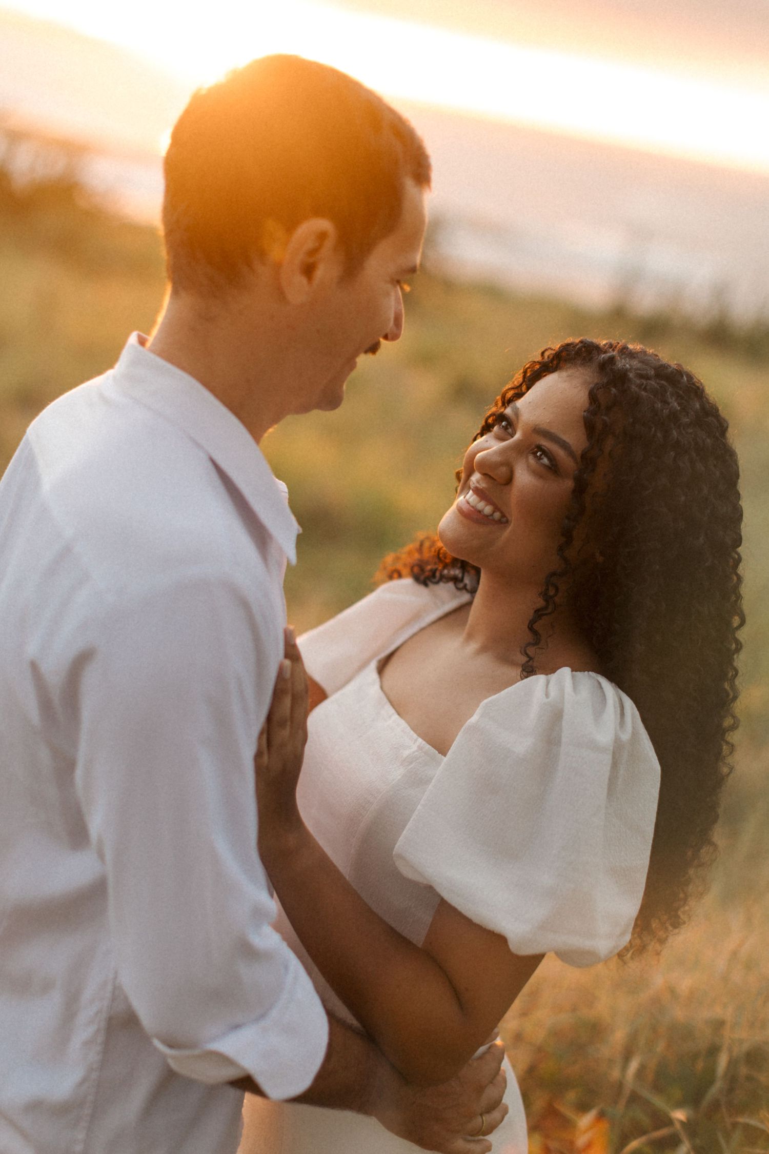 Romantic couple embraces in golden sunset light while standing in a grassy field wearing white clothing.