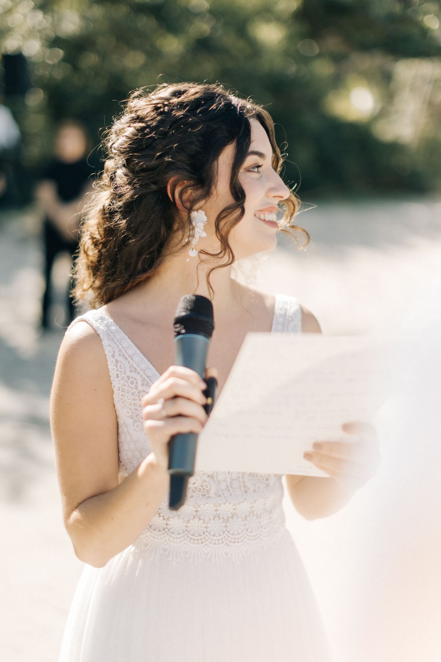 Bride in lace dress holding microphone and paper during outdoor wedding speech.