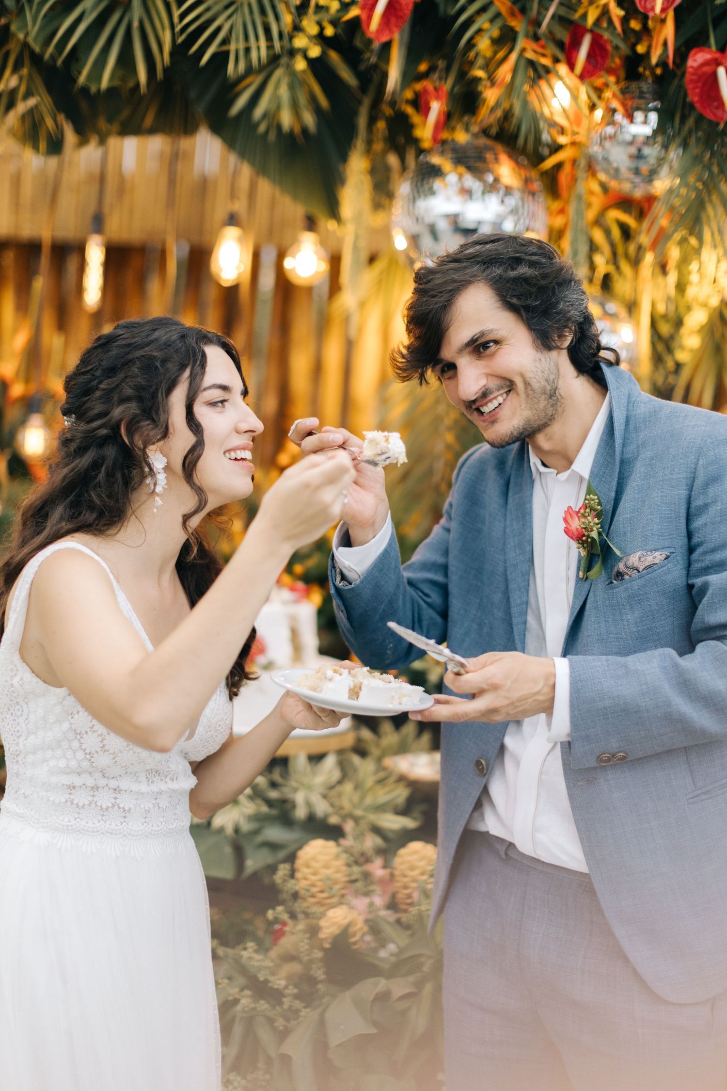 Newlyweds share cake at a reception decorated with palm leaves and hanging lights.