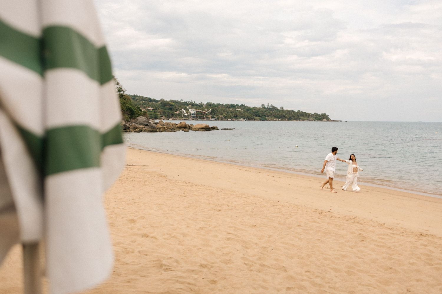 Two people in white outfits walk along a scenic tropical beach with turquoise water and green umbrellas visible.