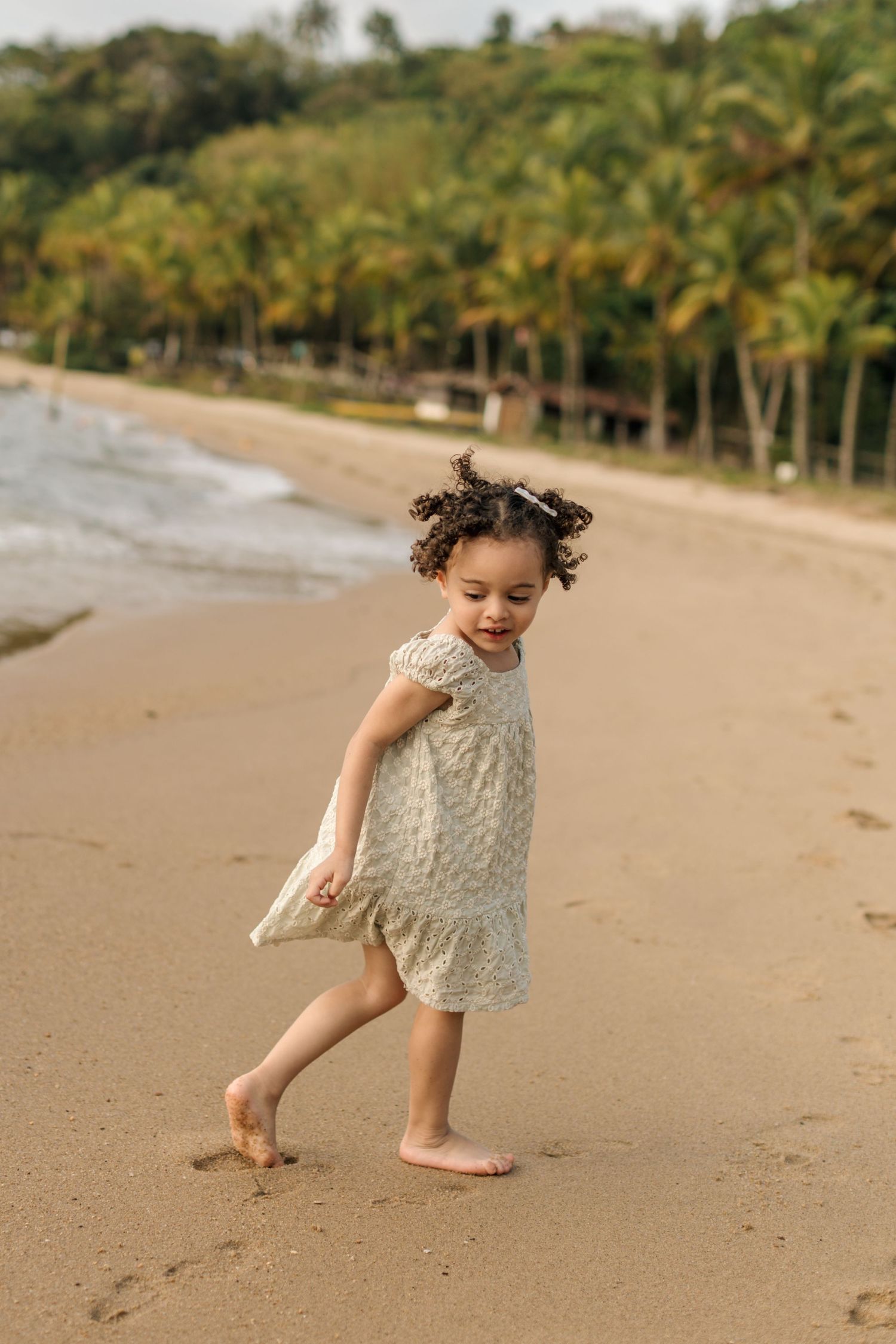 A child in a white lace dress plays joyfully on a tropical beach with palm trees in the background.