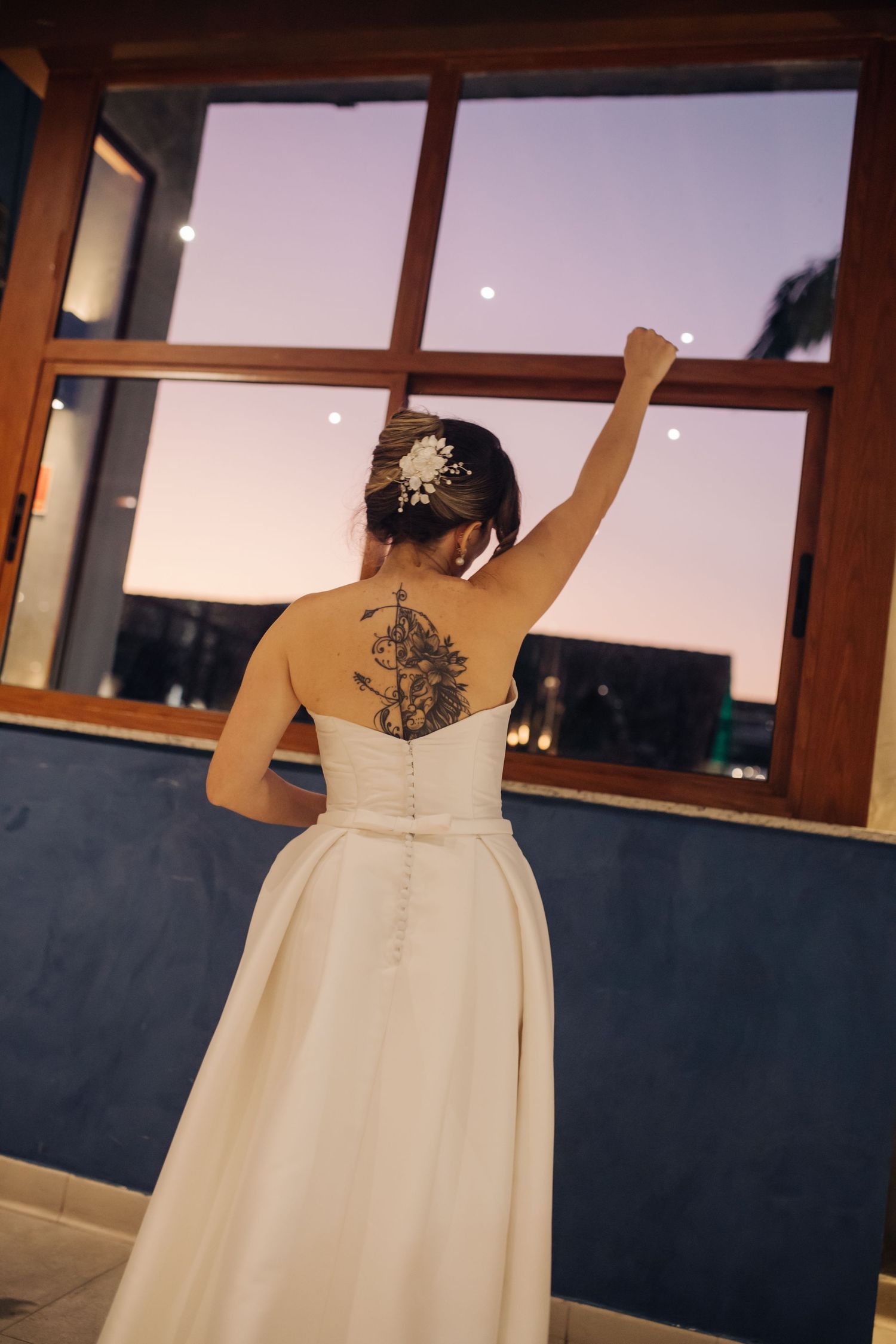 Back view of bride in white wedding dress showing intricate back tattoo and hair accessory against window.