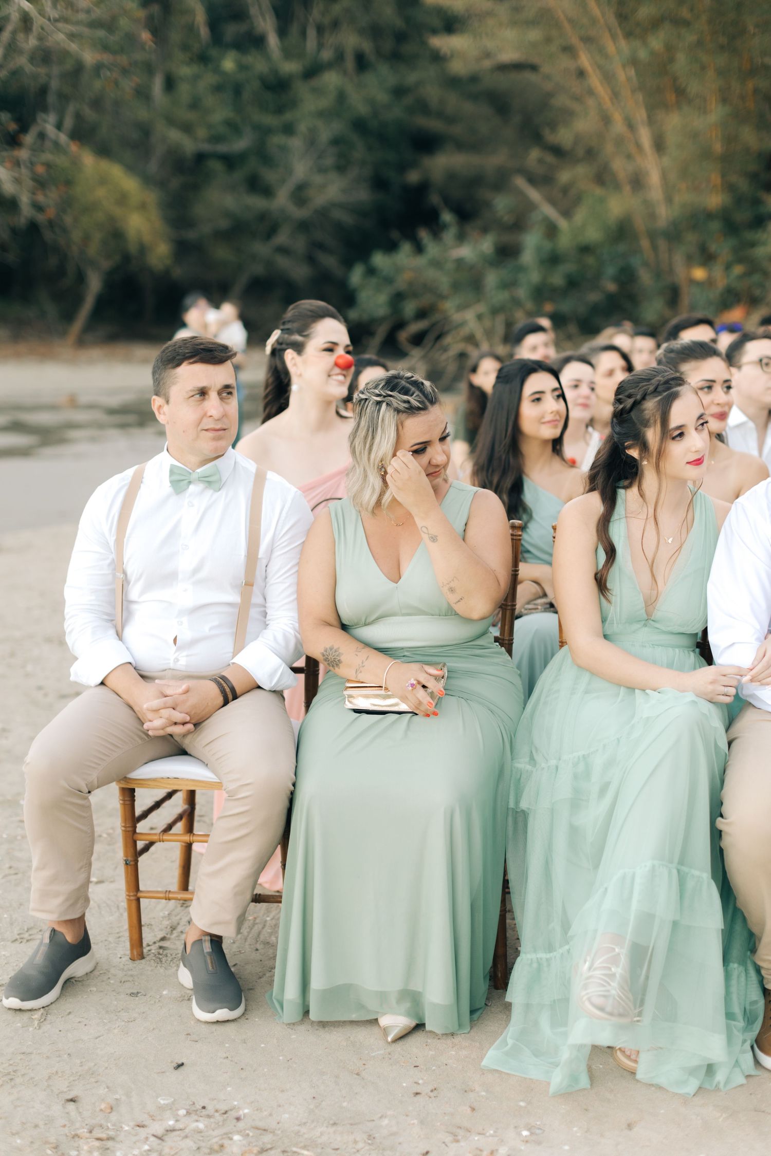 Wedding guests in mint green dresses and casual attire seated on wooden chairs at beach ceremony.