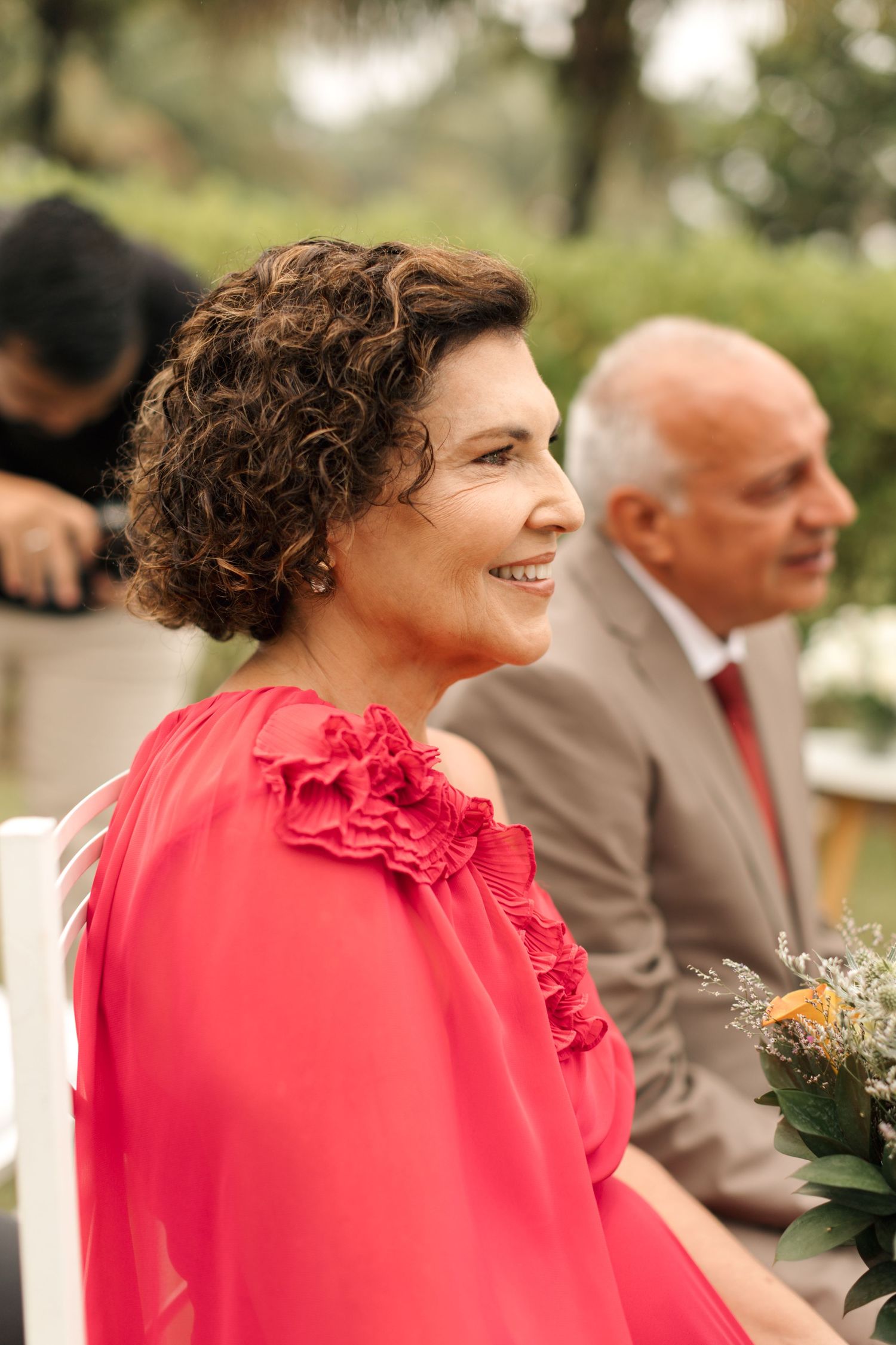 A woman in a bright coral dress with ruffled details smiles during an outdoor event while holding flowers.