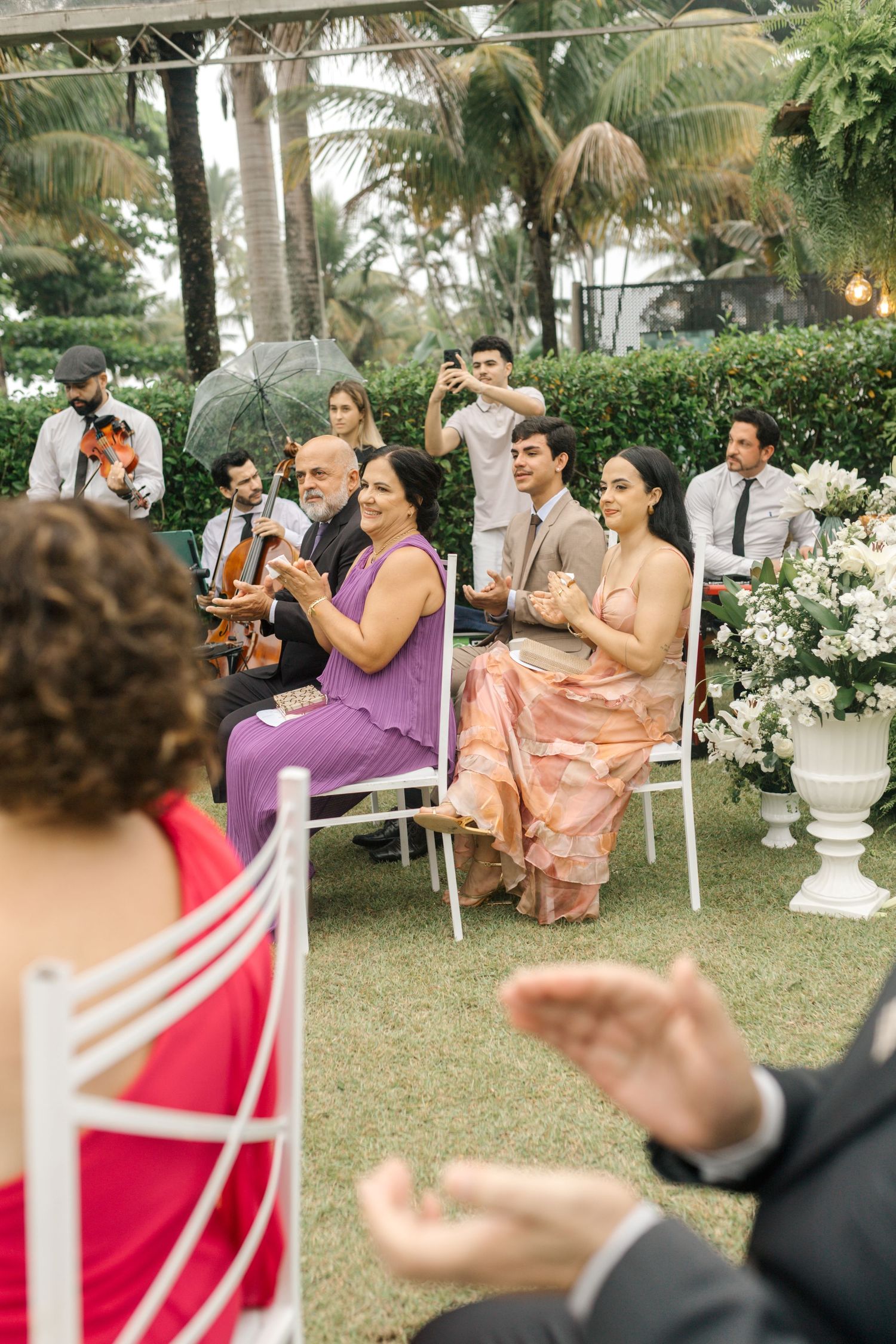 Wedding guests sitting in white chairs outdoors clapping during ceremony.