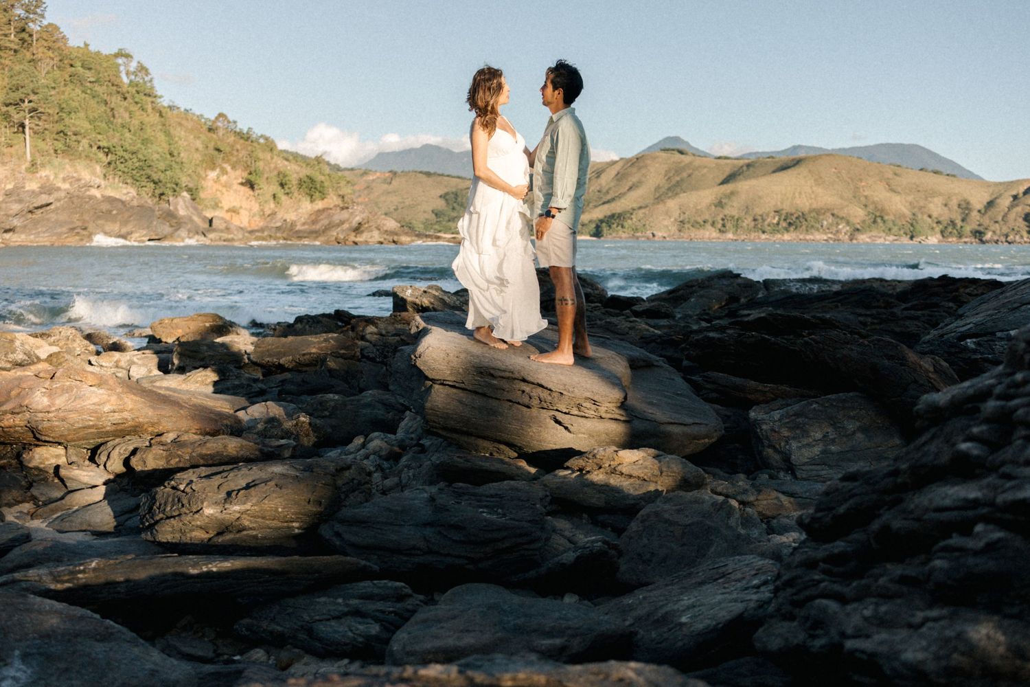 Two people stand on dramatic volcanic rocks at sunset overlooking a scenic coastal landscape.