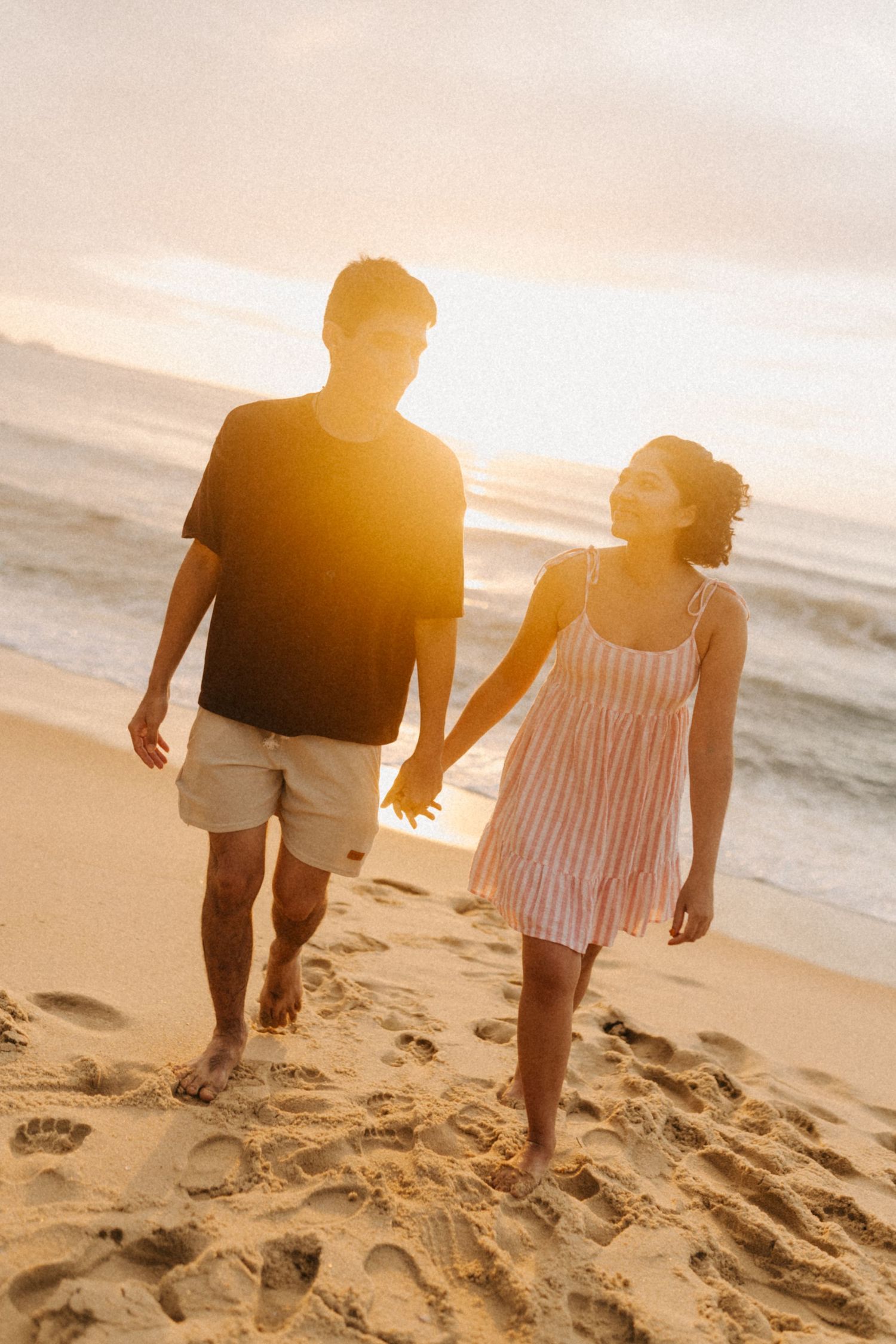Um casal caminha de mãos dadas em uma praia durante o pôr do sol da hora dourada com silhuetas quentes e iluminadas pelo sol.