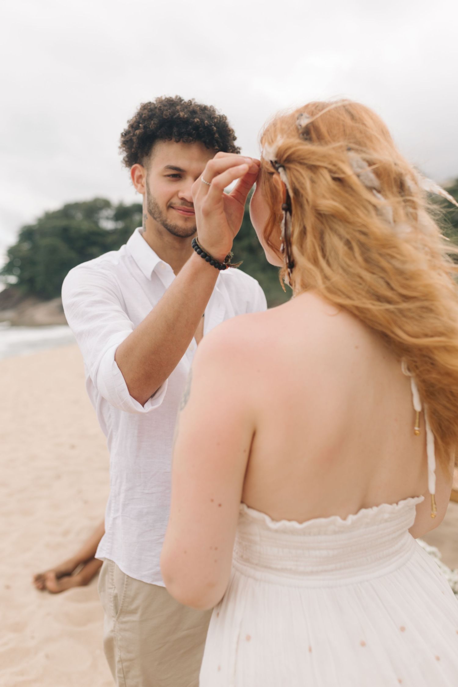Couple in white attire sharing an intimate moment on a beach during what appears to be a wedding ceremony.