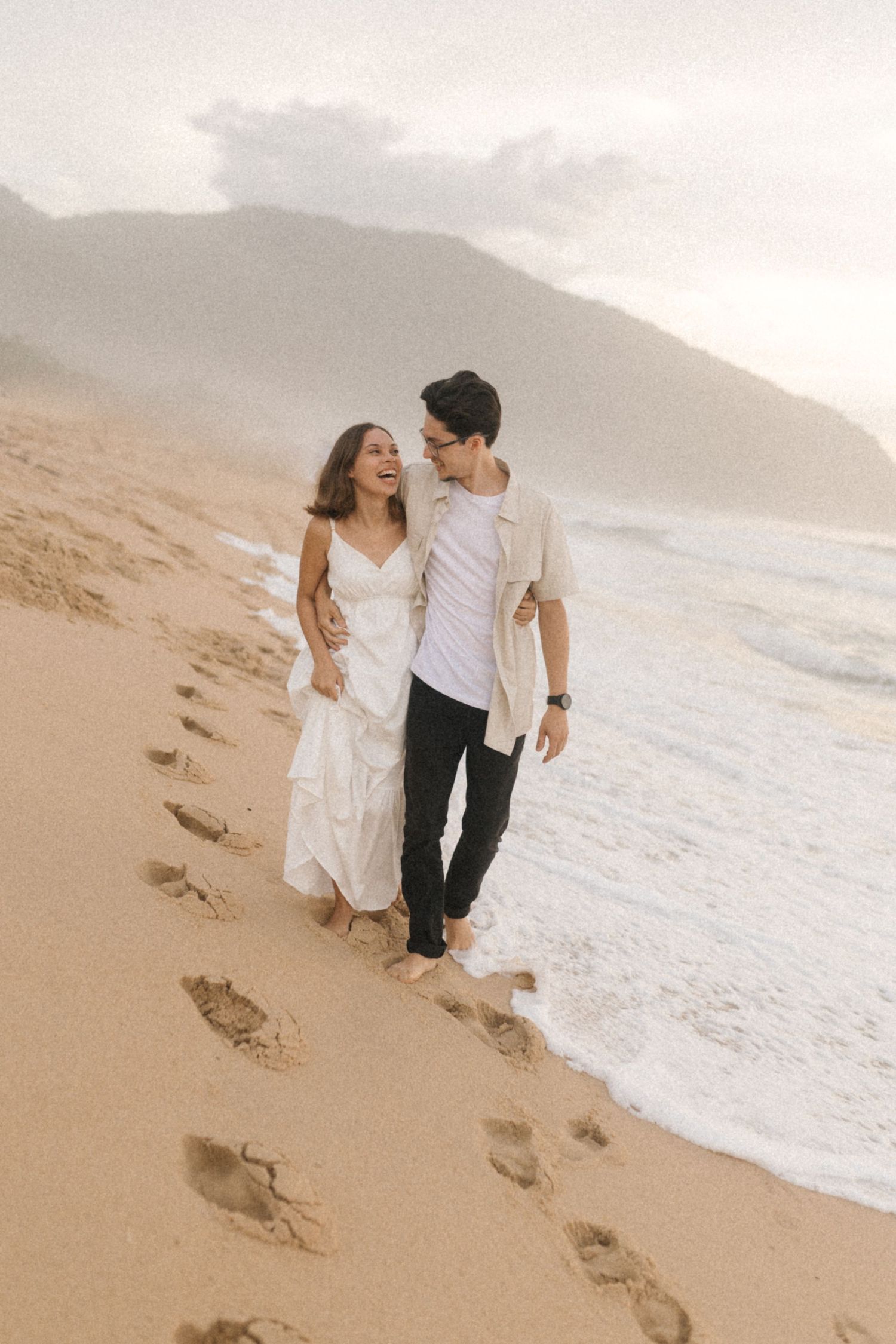 Romantic couple walking barefoot along a sandy beach with mountains and ocean waves in the background.