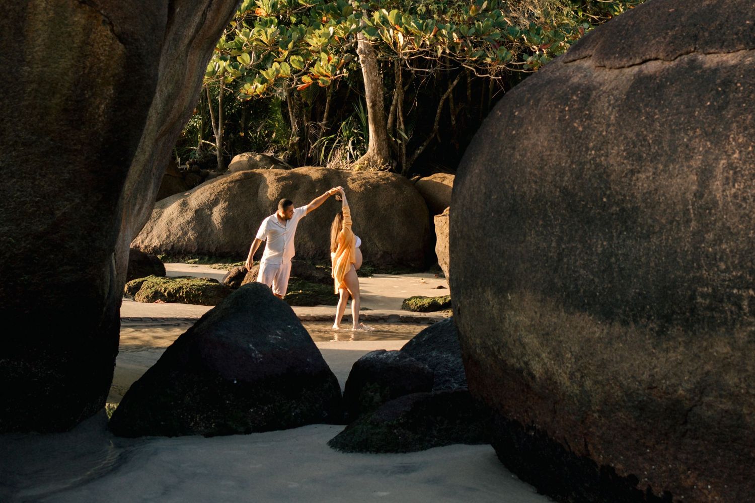 Silhouettes walking among large coastal boulders on sunny beach.
